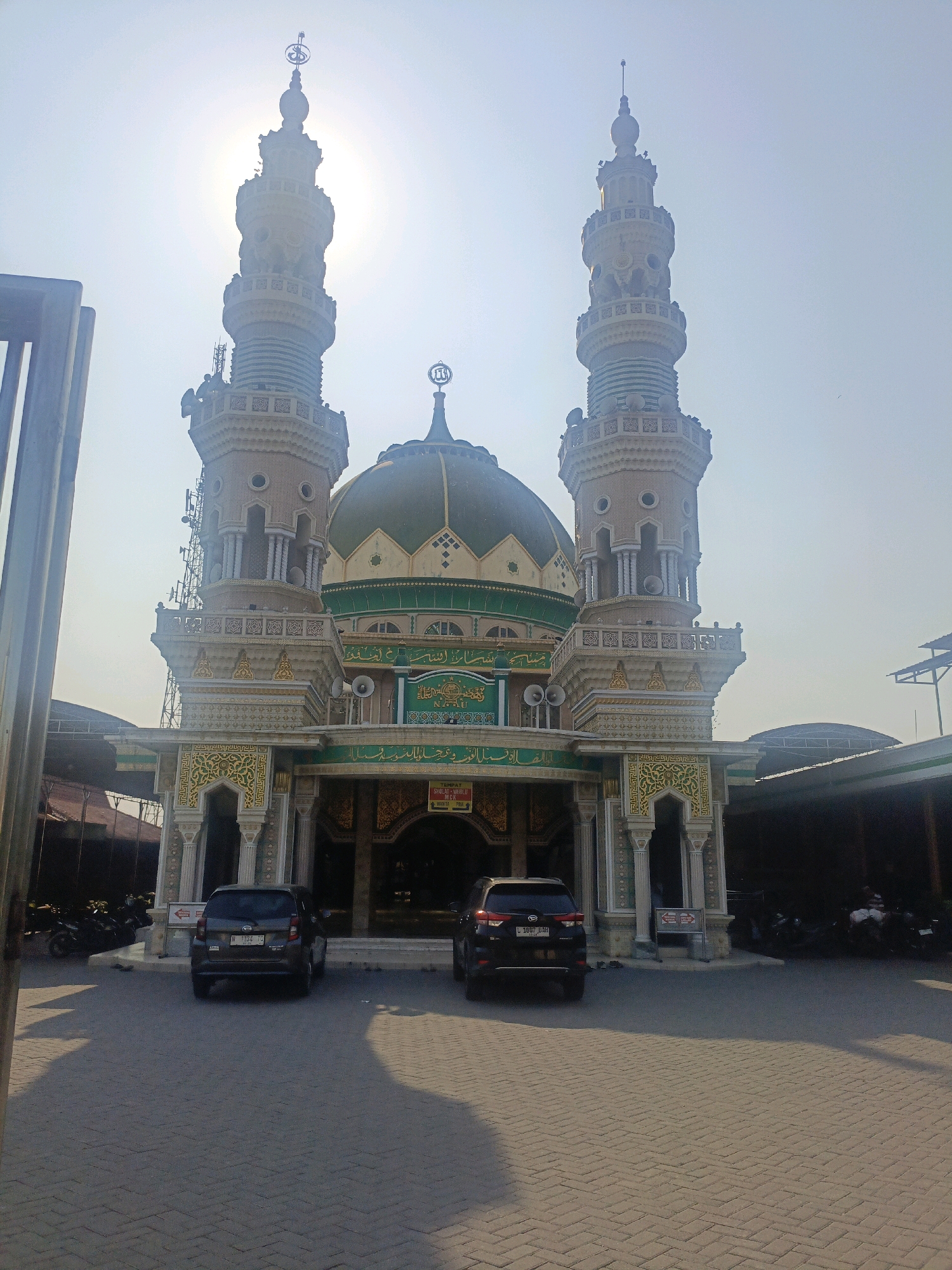 The image shows a mosque with two minarets and a dome, located in Gempol, Indonesia. The mosque is made of stone and has intricate carvings. In front of the mosque, there are two cars parked, a dark gray SUV and a black SUV. There are also several motorcycles parked to the left of the mosque. The sky is clear and the sun is shining. The area in front of the mosque is paved with cobblestones. The overall scene suggests a public gathering or a place of worship. The mosque appears to be well-maintained and is a prominent landmark in the area.
