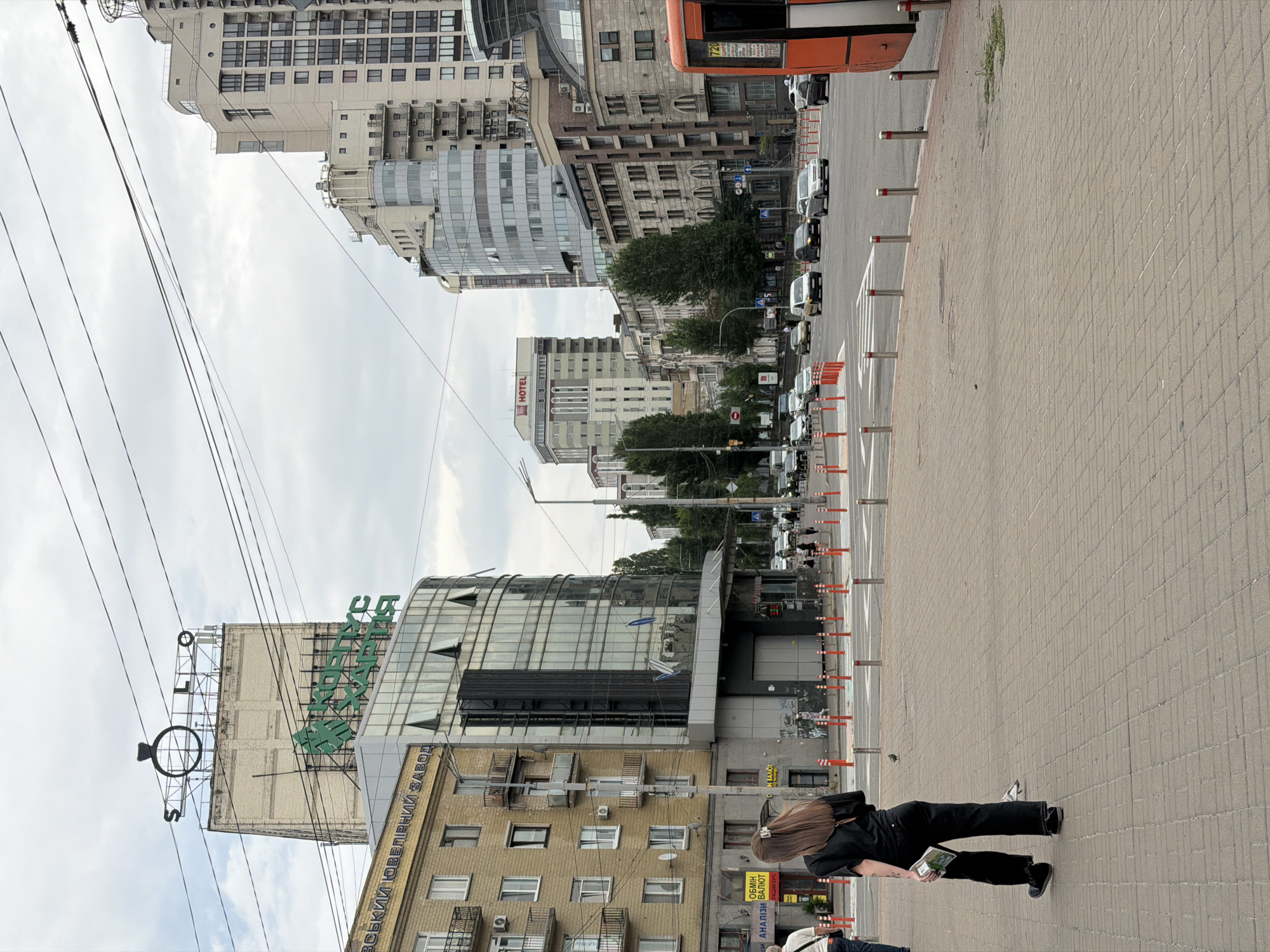 This is a street-level, full-shot image taken in Kyiv, Ukraine. The foreground shows a pedestrian sidewalk paved with light brown bricks, with a woman walking away from the camera towards the left side of the frame. She is wearing black pants and a black t-shirt, and carrying a small white object in her left hand. A few metal bollards are visible along the edge of the sidewalk.
The middle ground depicts a busy intersection with several cars driving in various directions. A crosswalk is marked on the road. On the left side of the intersection, there is a yellow brick building with balconies, and adjacent to it, a modern building with a glass facade. Above these buildings, signs are visible, including one with a large ring logo and the word "S.LO". Another green sign reads "КОРПУС ХАРТІЯ". Further down the street, a tall, modern hotel with an "IBIS HOTEL" sign is visible.
The background features a cluster of multi-story buildings, typical of an urban environment. The sky is overcast with grey clouds. On the right side of the frame, the rear of an orange bus or tram is partially visible. The overall scene captures a typical day in a city with a mix of older and modern architecture, and bustling street activity.