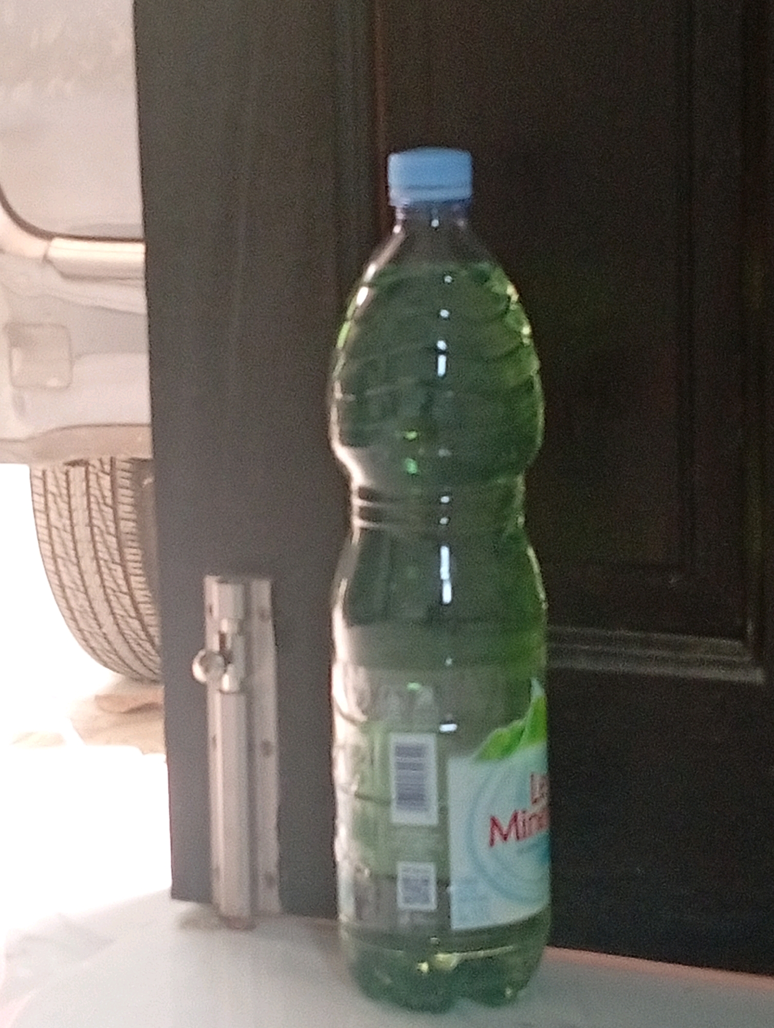 In the city of Samarinda, Indonesia, a clear plastic bottle filled with green liquid and a blue cap stands on a white surface. The bottle has a label that reads "Les Mina" in red letters, with a green leaf design. Behind the bottle, a dark brown door and a part of a white vehicle, including a tire, are visible. A silver latch or lock mechanism is attached to a dark surface to the left of the bottle. The scene appears to be indoors, with lighting suggesting it could be daytime.