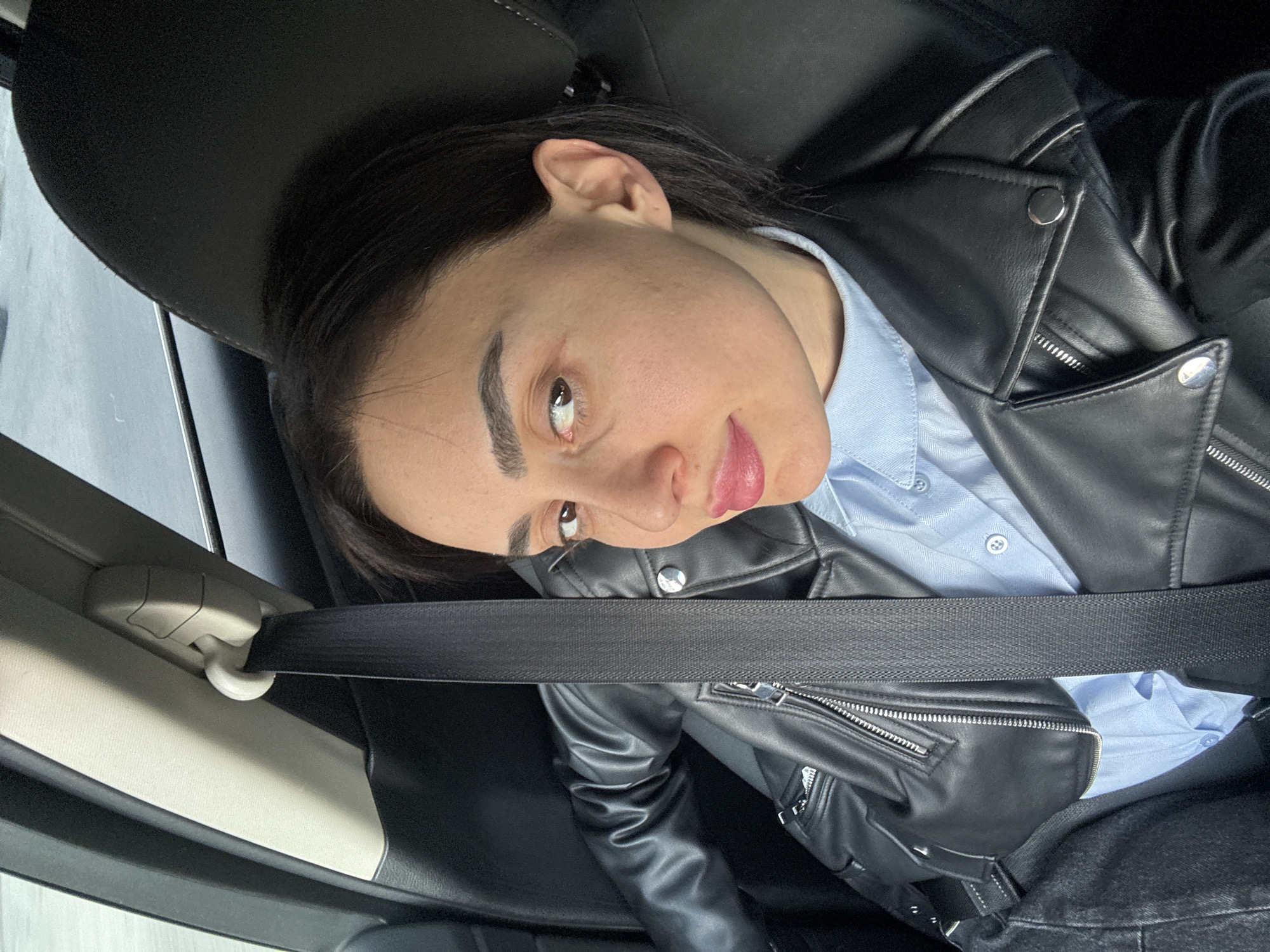 A person with brown eyes, eyebrows and hair is seated in a vehicle with a seatbelt fastened. They are wearing a light blue collared shirt and a black leather jacket. The interior of the car is black and grey. The location cannot be determined from the image.