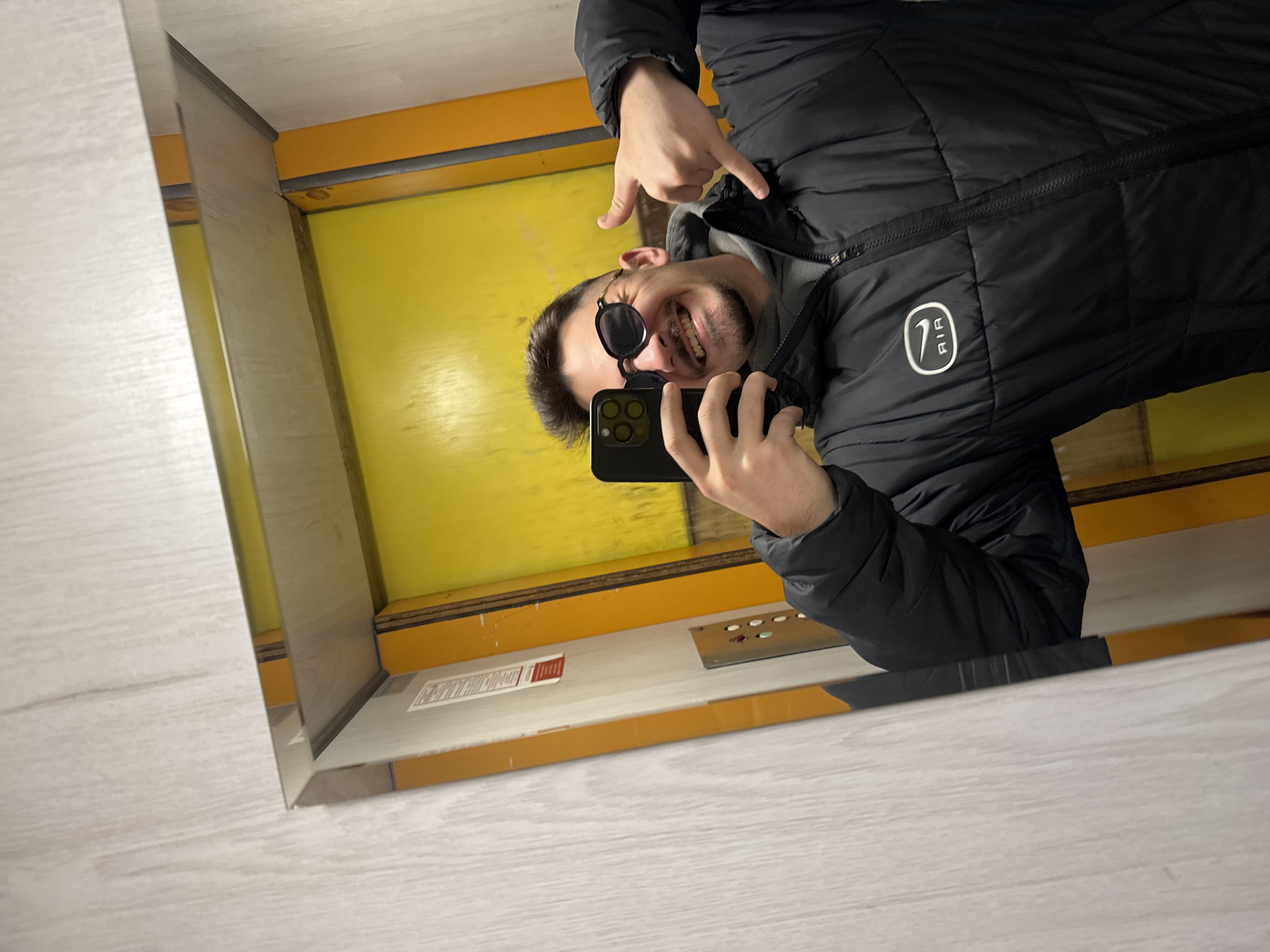 A man is taking a selfie in a mirror within an elevator with yellow walls. He is wearing a black puffer jacket and sunglasses. He has a short beard and is smiling. He is pointing down at himself with his left hand. He is holding a black smartphone in his right hand. The elevator's control panel is visible. The location is possibly in Sofia, Bulgaria, but there is no visual information to definitively confirm this.
