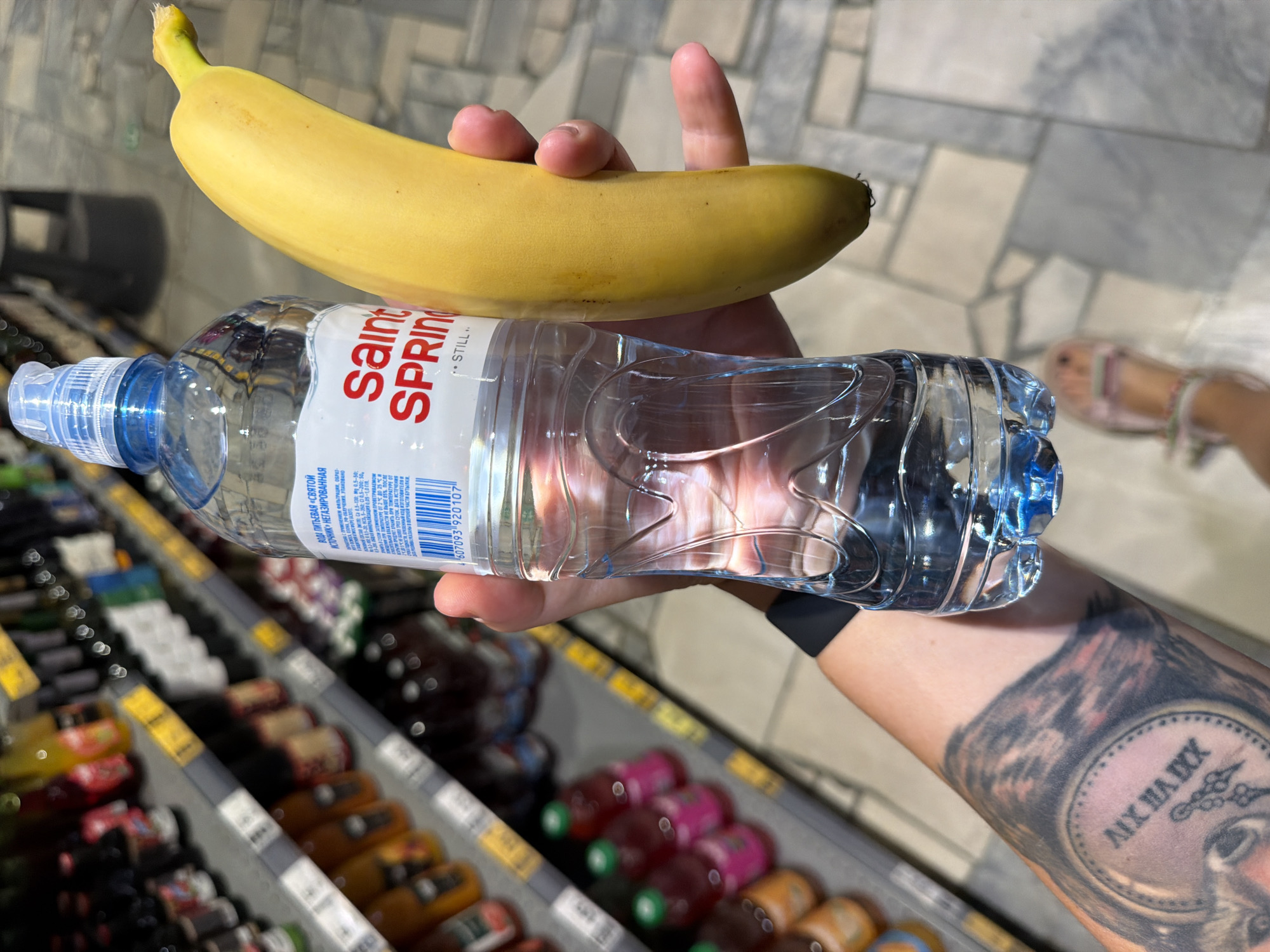 In a supermarket aisle, a person's arm and hand are visible, holding a clear plastic bottle of Saint Spring water and a ripe yellow banana. The water bottle has a blue cap and a label with red text. Behind them, shelves are stocked with various bottled beverages, including some with red liquid. The floor is tiled with a light grey and beige pattern. The person's arm displays a large tattoo of a clock and possibly an animal's face. The scene appears to be in the city of Chaikovsky, Russia, given the text on the water bottle label.