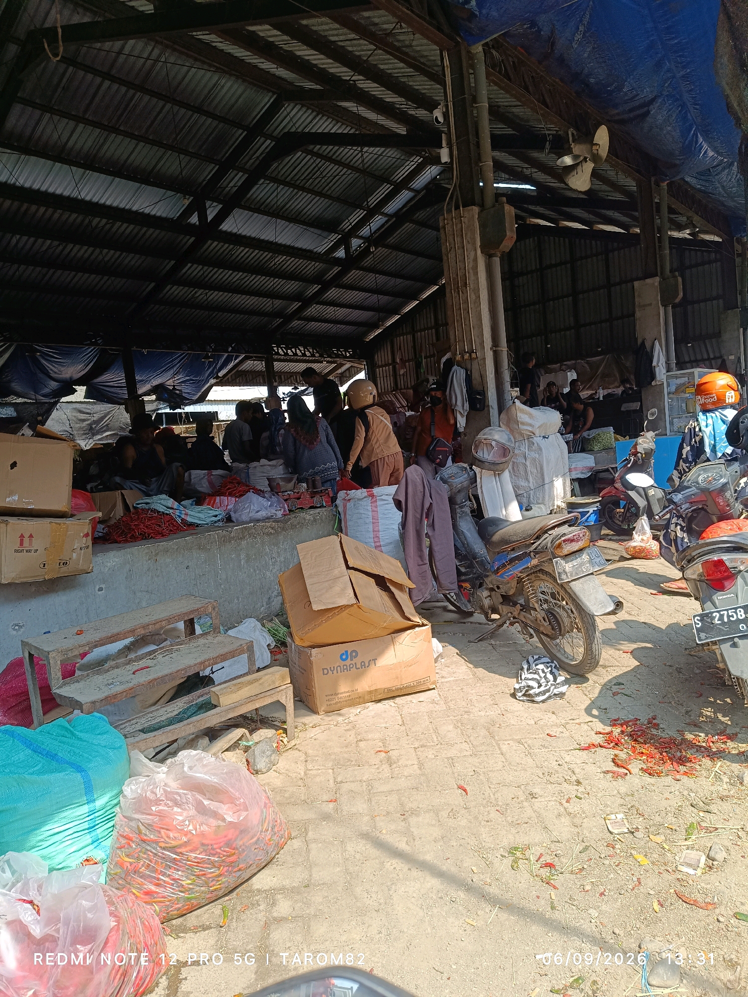 This image depicts an outdoor marketplace, likely a traditional market, in Pare, Indonesia. The scene is filled with various goods and activity. In the foreground, several large sacks are visible; one is a bright turquoise and appears to contain produce, while another is transparent and filled with red chili peppers. Next to these is a large, clear plastic bag also filled with red chili peppers. Several cardboard boxes are stacked, some with shipping labels. A wooden bench or makeshift seating area is present. In the mid-ground, several motorcycles are parked, some with helmets on their seats. A pile of red chili peppers is scattered on the ground near one of the motorcycles. Further back, under a large, open-sided structure with a corrugated metal roof and blue tarpaulin coverings, a number of people are gathered. They appear to be vendors and shoppers, with some individuals sitting and others standing or interacting with goods, which seem to include produce and other market items. The environment appears dusty and somewhat unorganized, typical of a bustling market. The overall impression is one of a busy commercial hub.