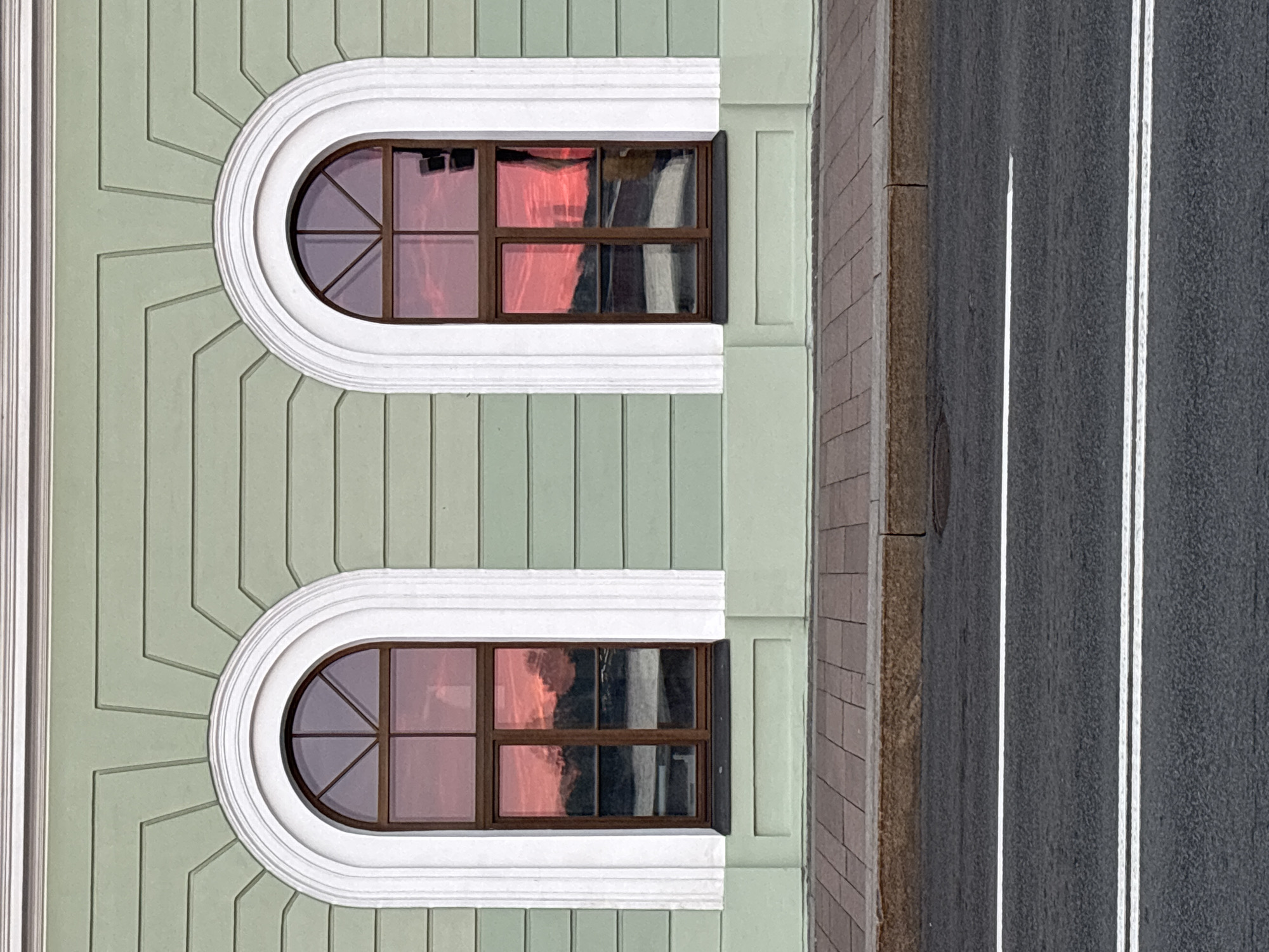 An elevated perspective captures a section of a building facade alongside a street in Saint Petersburg, Russia. The building, occupying the left portion of the image, features a light green exterior composed of vertical paneling. Two distinct, identically designed arched windows are prominent, each encased by ornate white moldings. These moldings create an elliptical frame around the upper section of the windows, with angular, ray-like patterns extending from them onto the green facade.
The windows have dark brown frames, and their glass brilliantly reflects a reddish-pink light, strongly suggesting either a sunrise or sunset. Faint, dark, indistinct shapes, possibly silhouettes of other buildings or trees, are visible within these reflections. The internal grilles of the windows divide the glass into sections, showcasing a fanned, semi-circular pattern within the arch and rectangular panes below.
To the right of the building, a dark grey asphalt road is visible, marked by two parallel white solid lines and a single broken white line. Adjacent to the road, a sidewalk constructed from reddish-brown bricks or pavers extends along the curb, where a portion of a manhole cover can be seen. The scene is static; no people or visible text are present.