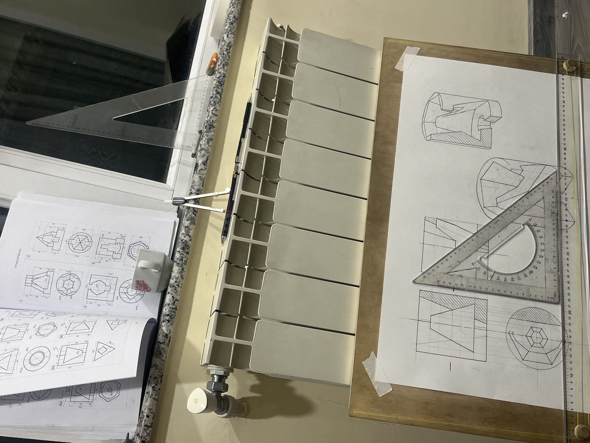 The image captures an indoor workspace dedicated to technical drawing, likely in Yerevan, Armenia. A wooden drawing board, positioned on the right, holds a large sheet of paper with intricate engineering or architectural drawings, depicting various geometric forms and cross-sections. A T-square and a protractor set square are neatly laid over the drawing.
To the left, an open textbook or binder displays similar technical diagrams, possibly exercises or examples. A small, white ceramic cup with a red pattern rests on this book. Adjacent to the book, a drawing compass is visible. Another transparent set square is placed on a granite-patterned window sill, which runs along the bottom of a window revealing a dark, ambiguous outdoor scene, suggesting it might be nighttime or a low-light environment. The room's wall is painted a light beige or yellow.
A modern, multi-section radiator is mounted on the wall between the drawing area and the window, with its control valve visible at the bottom. The overall impression is one of focused study or work.
Visible text includes Armenian words "9.Բարդ խնդիրներ" (meaning "9. Complex problems") on the open book, indicating it's a technical textbook or exercise guide. Various numbers, likely measurements or page/exercise numbers (e.g., 100, 0120, 89, 64, 3), are also scattered across the book's pages and ruler markings.