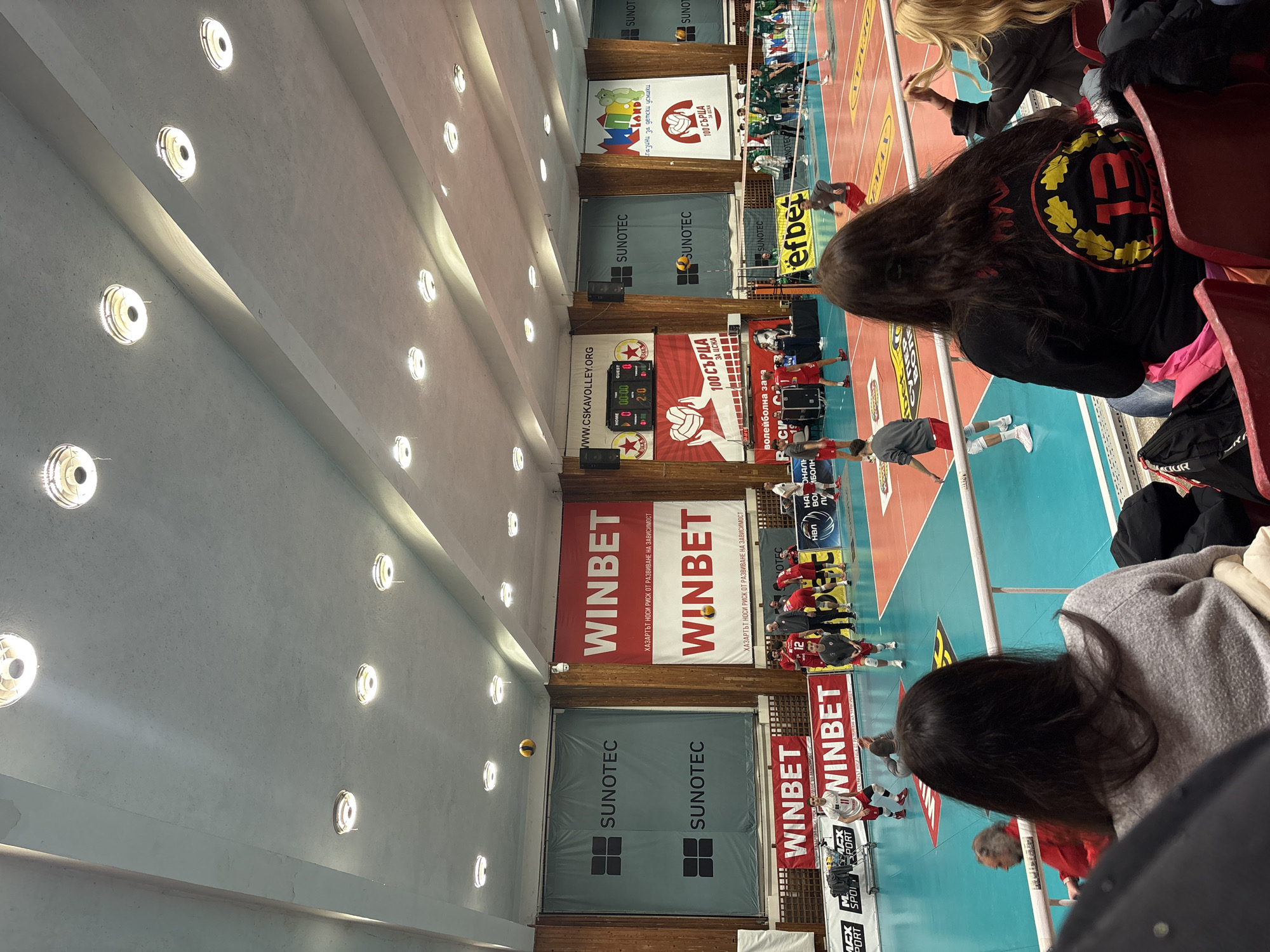 The image depicts an indoor volleyball match in progress at Vasil Simov Hall, located within the CSKA Sports Complex in Sofia, Bulgaria. The perspective is from the spectator stands, looking down towards the court.
The court features a blue and orange playing surface with white boundary lines. A net divides the court, and a yellow and blue volleyball is airborne above the net on the left side. Two teams are present: one wears red jerseys with white numbers (e.g., player #12 visible, another player in a receiving stance), and the opposing team wears dark green or black jerseys. Players are actively engaged in play or awaiting action.
In the foreground, the backs of two female spectators are visible, seated in red stadium seats behind a white railing. One woman wears a gray sweater, and the other wears a black top with a prominent circular yellow and red emblem on the back, featuring the number "13" within a laurel wreath. Other spectators are partially visible further down the row.
The walls of the hall are paneled with wood in the lower sections, with large advertising banners above. Prominent banners include multiple red and white "WINBET" advertisements (some stating "ХАЗАРТЪТ НОСИ РИСК ОТ РАЗВИВАНЕ НА ЗАВИСИМОСТ" - Gambling carries a risk of addiction) and teal/grey "SUNOTEC" banners. A scoreboard on the left wall displays "HOME 0", "GUEST 0", and "2:0", with "WWW.CSKAVOLLEY.ORG" and a "100 СЪРЦА ЗА ЦСКА" (100 Hearts for CSKA) banner beneath it, featuring a volleyball logo. An "efbet" logo is visible on a mat on the court, and a "KIDS LAND" banner with "Магазини за детски усмивки" is seen on the right. Security cameras and speakers are mounted on the upper walls.
The ceiling is high, light-colored, and features multiple rows of circular, recessed light fixtures illuminating the hall.