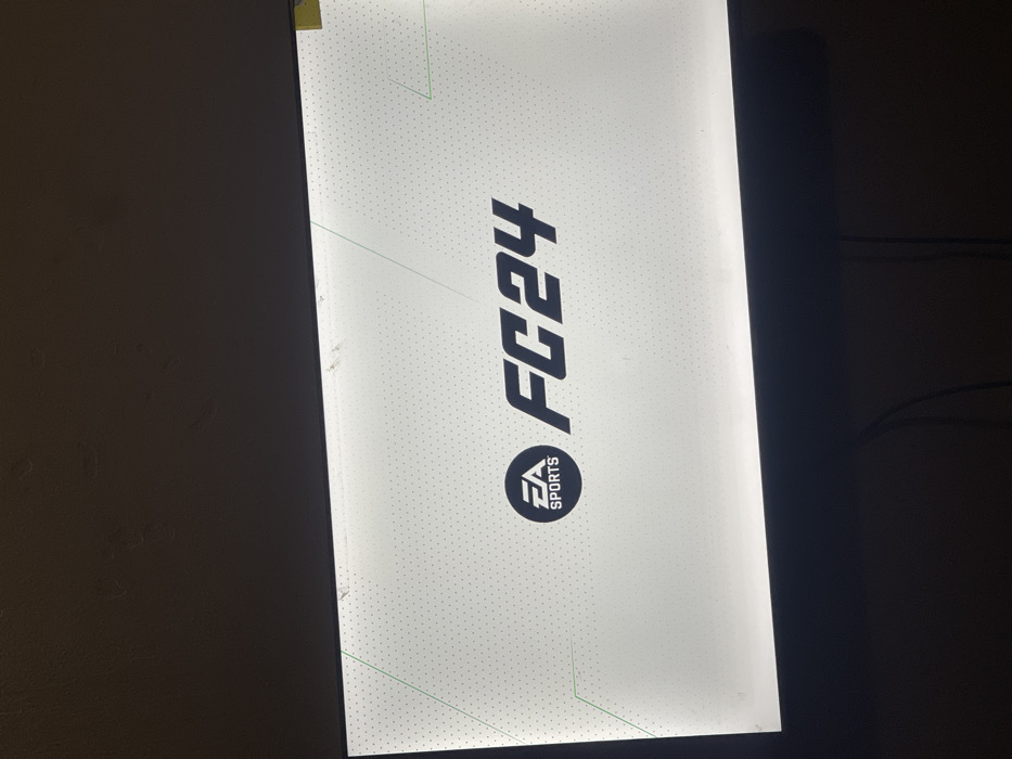 An illuminated sign displays "FC24" in a bold, slanted font above the EA Sports logo. The background is white with a subtle dotted pattern and faint green accents in the corners. The sign appears to be rectangular and mounted on a wall, which is dark. Visible smudges and imperfections can be seen near the edges of the sign. Wires are visible on the right.