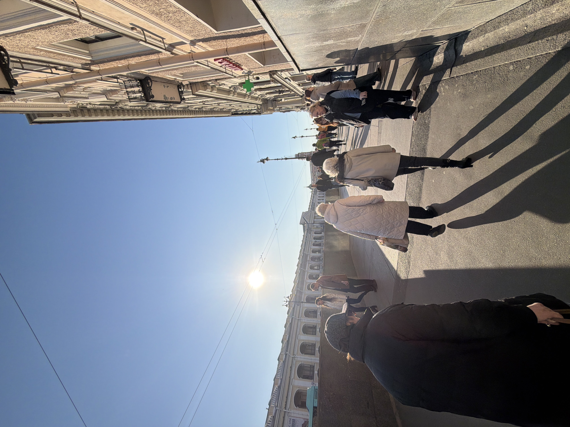 An urban street scene unfolds on a bright, sunny day in Saint Petersburg, Russia, specifically on Nevsky Prospekt, with the Bolshoy Gostiny Dvor building visible on the left. The clear blue sky is punctuated by overhead power lines.
On the left side of the wide street, the lengthy, two-story yellow facade of the Bolshoy Gostiny Dvor features numerous arched windows with white trim and a white balustrade along its roofline. On the right, a multi-story historical building with a textured, light-colored facade, ornate decorative elements, and black wrought-iron balconies lines the sidewalk. A prominent green cross pharmacy sign, with red Cyrillic lettering above it (likely "Аптека"), is affixed to this building.
The paved sidewalk in the foreground and midground is occupied by approximately twelve pedestrians, casting long shadows to their right, indicative of a low sun angle. In the immediate foreground, a person in a black puffer jacket and dark knitted hat walks away from the camera, holding a brown strap or handle. Ahead of them, two women also walk away; one wears a quilted light-colored coat, dark pants, and flat shoes, carrying a light bag; the other wears a light coat, dark pants, and black heeled boots, carrying a dark patterned handbag.
In the midground, several individuals move in both directions. One woman in a dark vest over a lighter top holds a smartphone as she walks towards the camera. Further back, a man in a brown jacket walks alongside a woman in a white jacket. A streetlamp pole is positioned in the midground, and various other pedestrians are visible in the distance, all dressed in winter or cool-weather attire. The scene depicts typical pedestrian activity on a major thoroughfare.