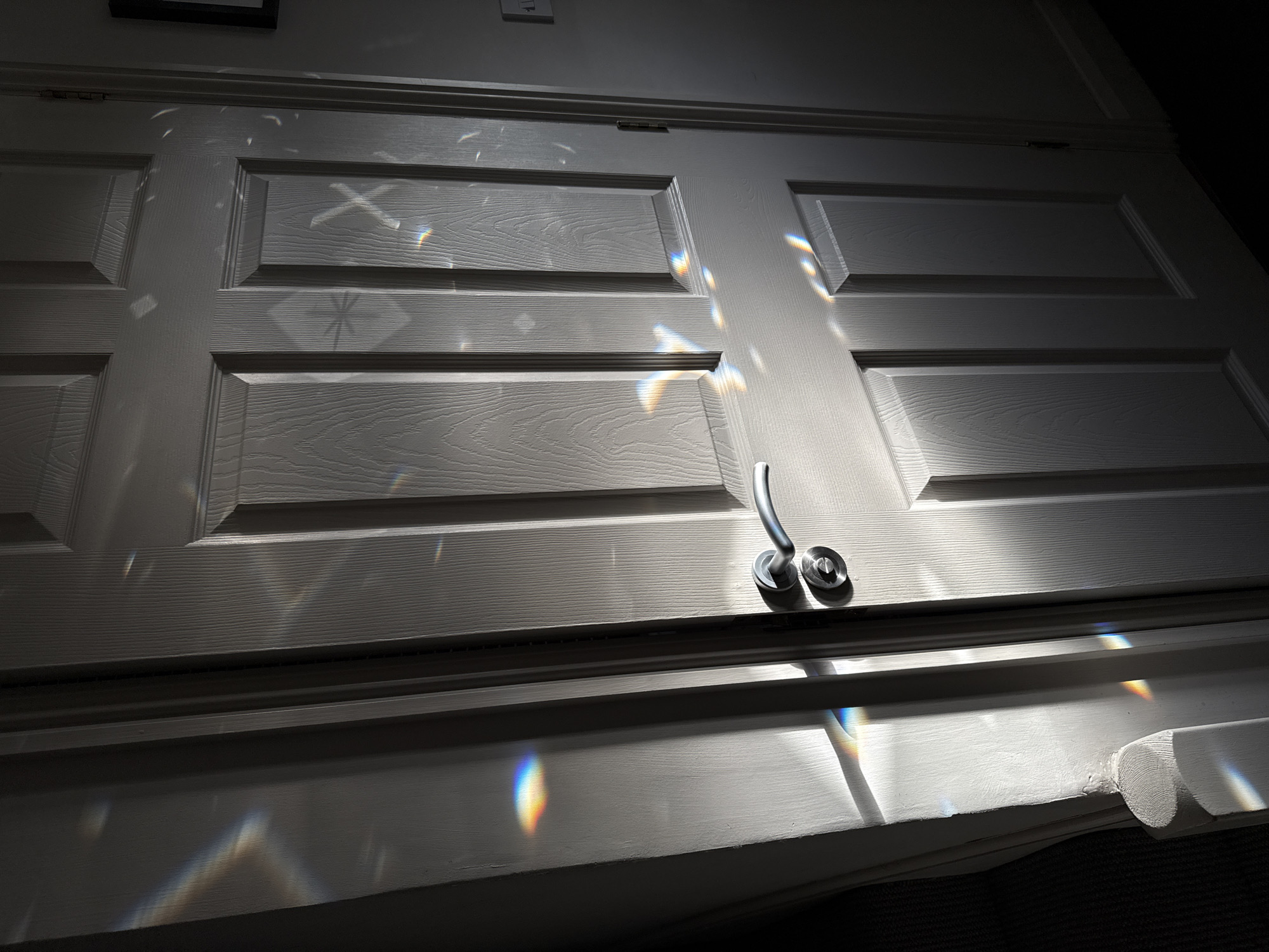 A low-angle shot depicts a white, paneled door with a silver handle and lock. Sunlight refracting through a prism casts rainbow-colored highlights across the door's surface. The door is likely located indoors. The image does not provide evidence for its location within East Suffolk, United Kingdom. There are no people present.