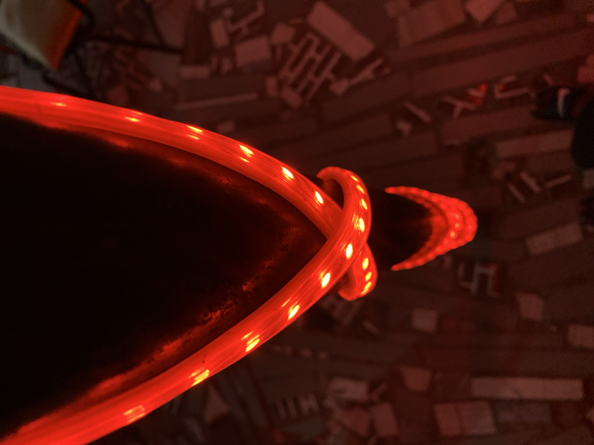 A close-up shot depicts a dark, possibly black, curved surface edged with a bright red LED strip. The LED strip is coiled around the edge of the surface, emitting a strong red glow. The background is dimly lit and appears to be a rough surface with various small, indistinct objects scattered across it. The color palette is dominated by reds and blacks, creating a high-contrast image. The location of the image in Kabong, Nigeria, is not discernible from the visual information.