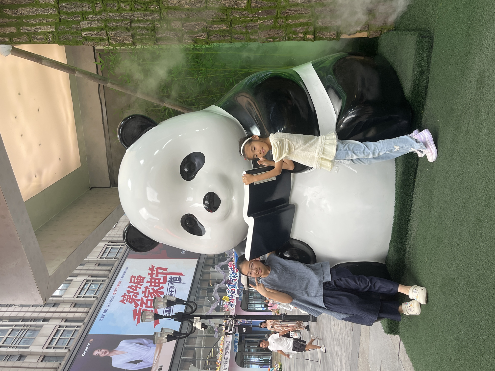 A full, medium shot captures two young girls posing with a large, glossy panda statue in an outdoor urban setting. The panda, depicted sitting and reading a book, is the central focus. The girl on the left, wearing a blue tunic and dark pants, leans against the panda's arm, holding up a peace sign. The girl on the right, in a white ruffled shirt and light blue jeans adorned with butterflies, stands beside the panda, also offering a peace sign. Both girls wear casual footwear. The background reveals a bustling city street with buildings, billboards advertising products, and pedestrian traffic, suggesting a commercial district. The overall scene is illuminated by daylight. The environment appears to be a public square or walkway, with the panda statue placed on an artificial grassy area.