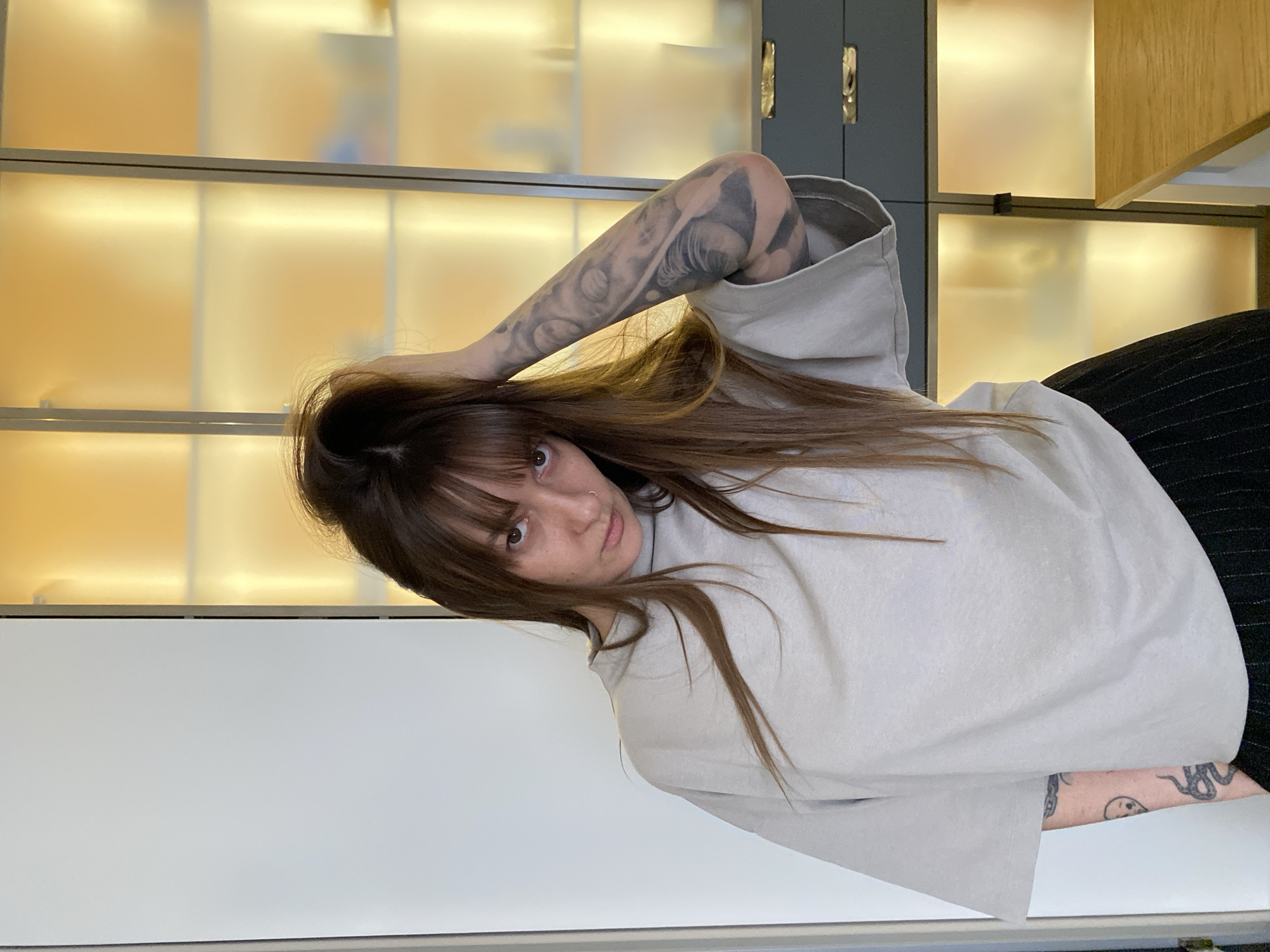 A woman stands indoors, possibly in an office or a professional setting. She has long brown hair with bangs and a nose ring. The woman is wearing a light grey t-shirt and a dark skirt. Her left arm is raised, and it has a tattoo on it. Her right arm also has a tattoo. Behind her is a wall with cabinets that are lit from the inside. The wall to her left is white. The image does not provide enough information to confirm its location in Votkinsk, Russia.