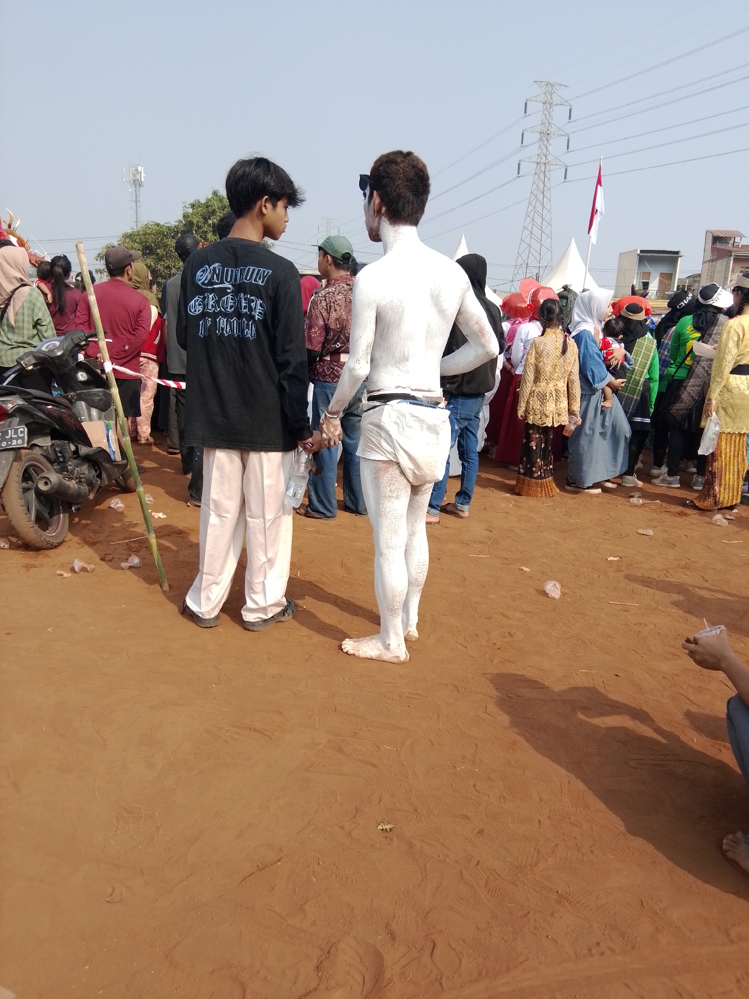 In the city of Bencongan, Indonesia, two young men are the central focus of an outdoor gathering. One man, on the left, wears a black long-sleeved shirt with white gothic-style lettering that reads "ONLY GROVES IF FOLK." He is also wearing loose-fitting white trousers and black shoes. He holds a clear plastic bottle of water in his left hand and a long bamboo pole in his right, which rests on the dusty, reddish-brown ground. The second man, on the right, is entirely covered in white paint, from his head to his feet, appearing as a statue. He wears only white briefs. Both men are facing away from the camera, looking towards a crowd of people in the background.
The crowd consists of individuals in various attire, suggesting a festive or community event. Some are wearing traditional Indonesian clothing, while others are in more casual wear. The background reveals a bright, clear sky, a few scattered trees, a large electrical tower with power lines, and several white tents. A red and white Indonesian flag is visible flying from a pole. A motorcycle is parked on the left side of the frame. The ground is covered in fine, reddish-brown dirt with scattered plastic debris. The overall scene suggests a public event taking place during the daytime under a sunny sky.