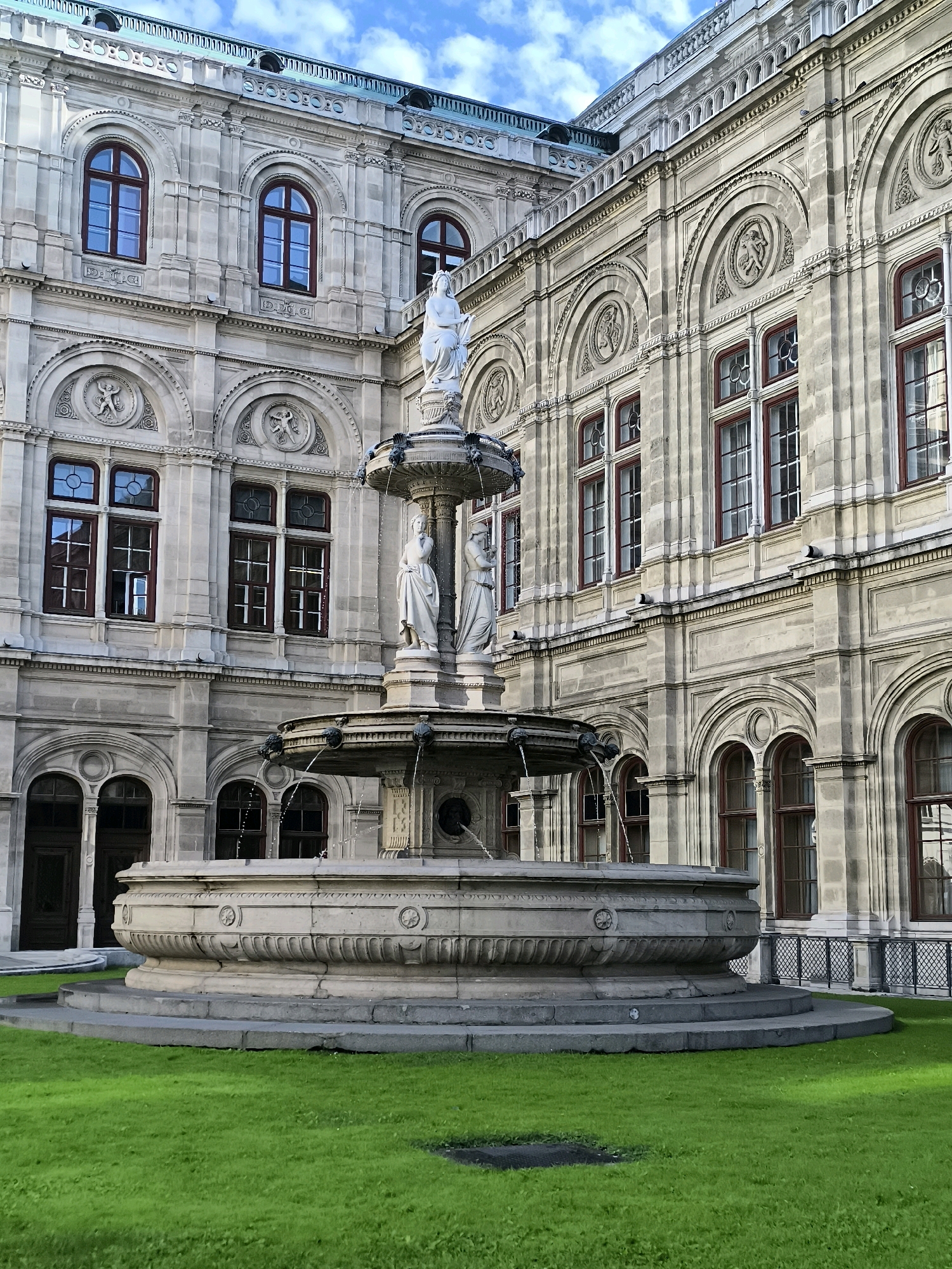 The image shows a view of the Vienna State Opera building in Vienna, Austria, with a prominent fountain in the foreground. The building is an imposing Neorenaissance structure made of light-colored stone, characterized by its ornate facade, arched windows with dark frames, and decorative architectural details such as sculpted reliefs and cornices. A large, tiered stone fountain with sculptures of human figures stands in the center of the frame, with water flowing from its upper levels. The fountain is surrounded by a neatly manicured green lawn. The sky above is partly cloudy.