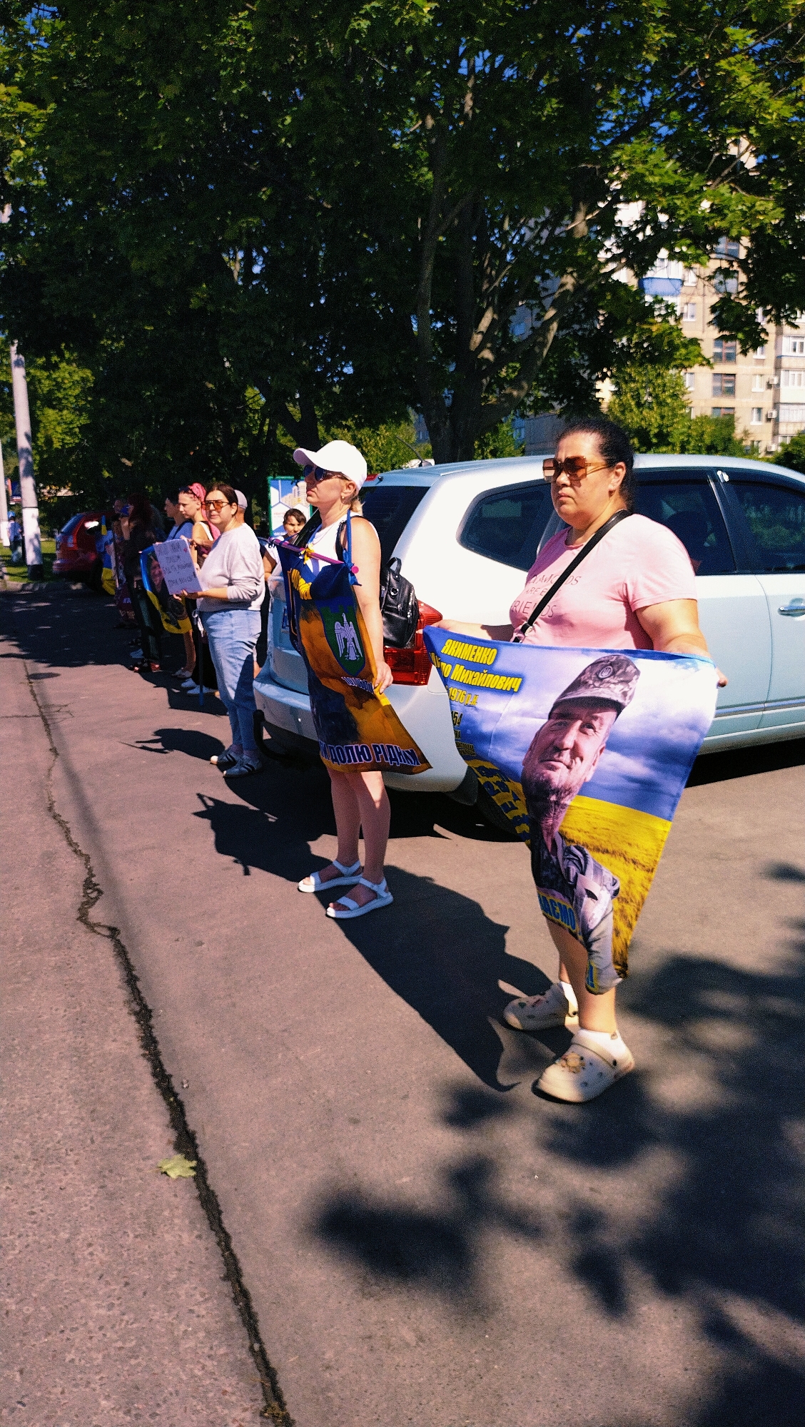 In Horishni Plavni, Ukraine, a group of individuals stand in a line on a paved street on a sunny day, casting long shadows from the trees overhead. They are holding several Ukrainian flags and posters. In the foreground, a woman in a pink t-shirt and sunglasses holds a large poster that features the portrait of a man in a military uniform against a backdrop of a yellow field and blue sky, with Ukrainian text printed on it. Another woman, to her left, wears a white cap and a white tank top, holding a flag with similar imagery. Further down the line, other individuals are visible holding signs and flags. A light-colored car is parked behind the people. The scene appears to be a demonstration or public gathering.