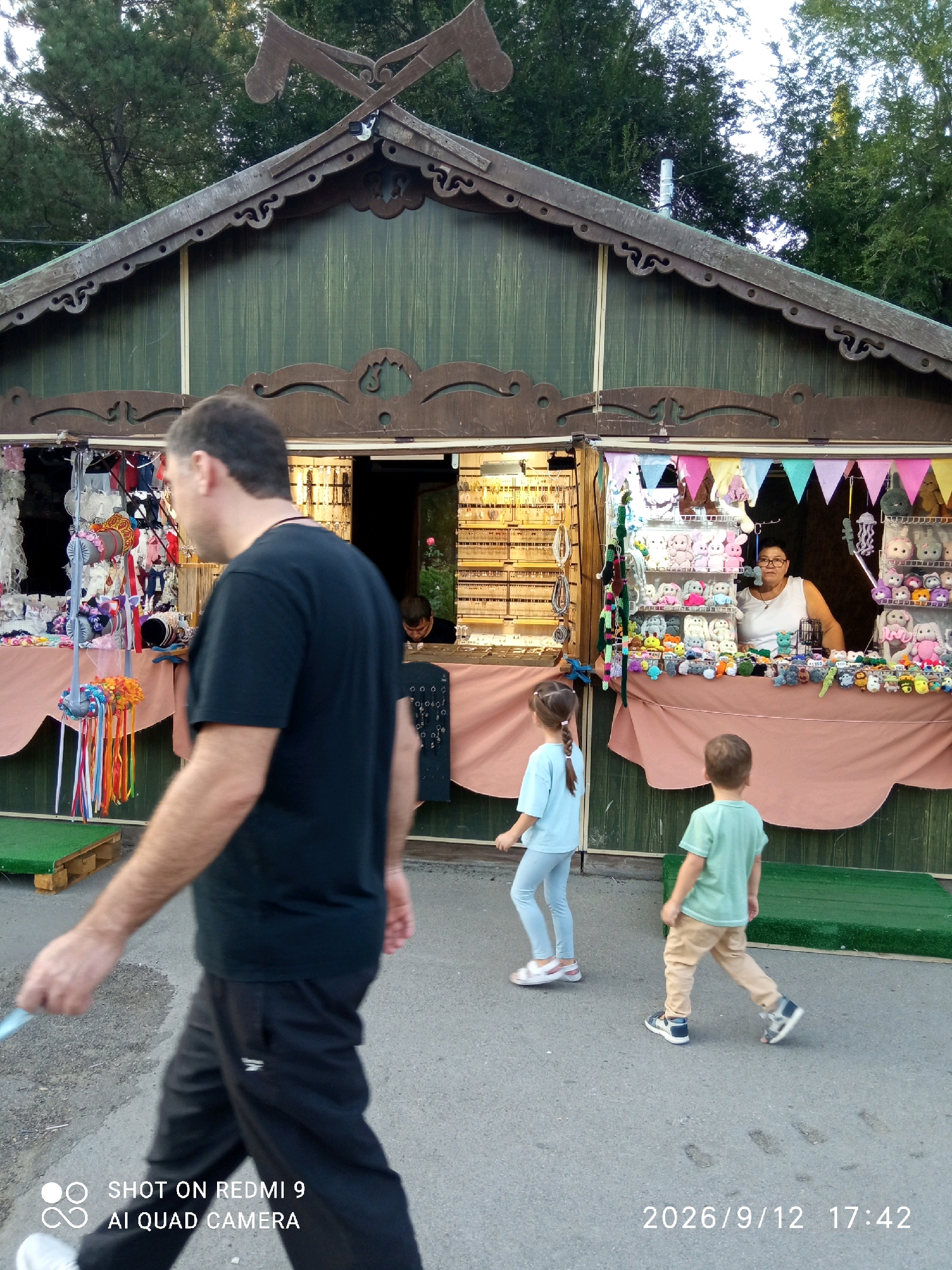 In the city of Rostov-on-Don, Russia, a scene unfolds at an outdoor market stall. The stall, constructed from dark wood with intricate carvings and a green wooden facade, displays a variety of goods. On the left, an assortment of colorful ribbons and trinkets hangs from a display rack. To the right, the stall is stocked with shelves filled with plush toys, many of which appear to be stuffed animals. A woman with glasses, wearing a white top, stands behind the counter, attending to the stall. In the foreground, a man with short dark hair, dressed in a black t-shirt and dark pants, walks away from the viewer, his left hand holding a light-colored object. Behind him, a young boy in a light green t-shirt and tan pants walks to the right, his gaze directed away from the camera. To his left, a young girl with a braid, wearing a light blue top and light blue leggings, walks in the same direction, her head turned slightly. The ground is paved asphalt, with faint footprints visible. The stall is situated outdoors, with trees and a clear sky visible in the background. A date and time stamp, "2026/9/12 17:42," is present in the bottom right corner of the image.