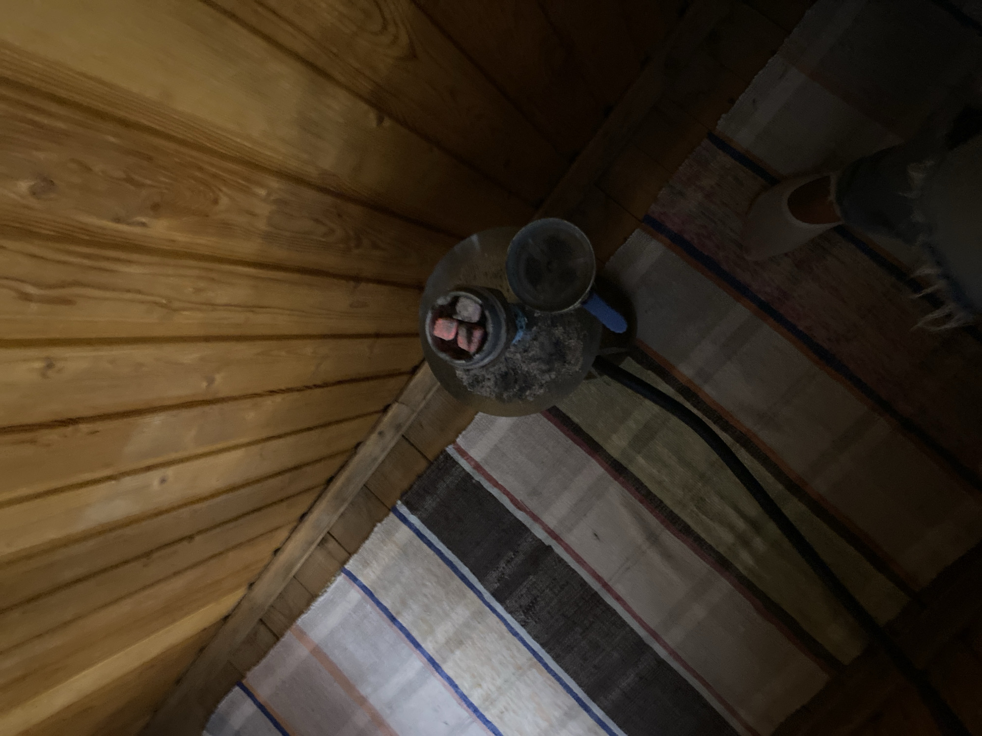 The image is a top-down shot of a hookah set up on a rug. The scene is indoors, with wooden walls and ceiling panels suggesting a rustic or cozy setting. A person's legs and feet, clad in a white slipper and jeans, are visible on the right side of the frame, indicating their presence in the scene. The rug beneath the hookah has a striped pattern with various colors including white, beige, brown, and blue. The hookah itself has a clear base filled with ash and water, a metal stem, and a bowl filled with red-hot coals and dark shisha tobacco. The lighting is dim, suggesting it might be evening or a dimly lit room. The overall atmosphere appears relaxed and conducive to smoking a hookah. There is no visible text in the image. The context provided places this scene in Perm, Russia.