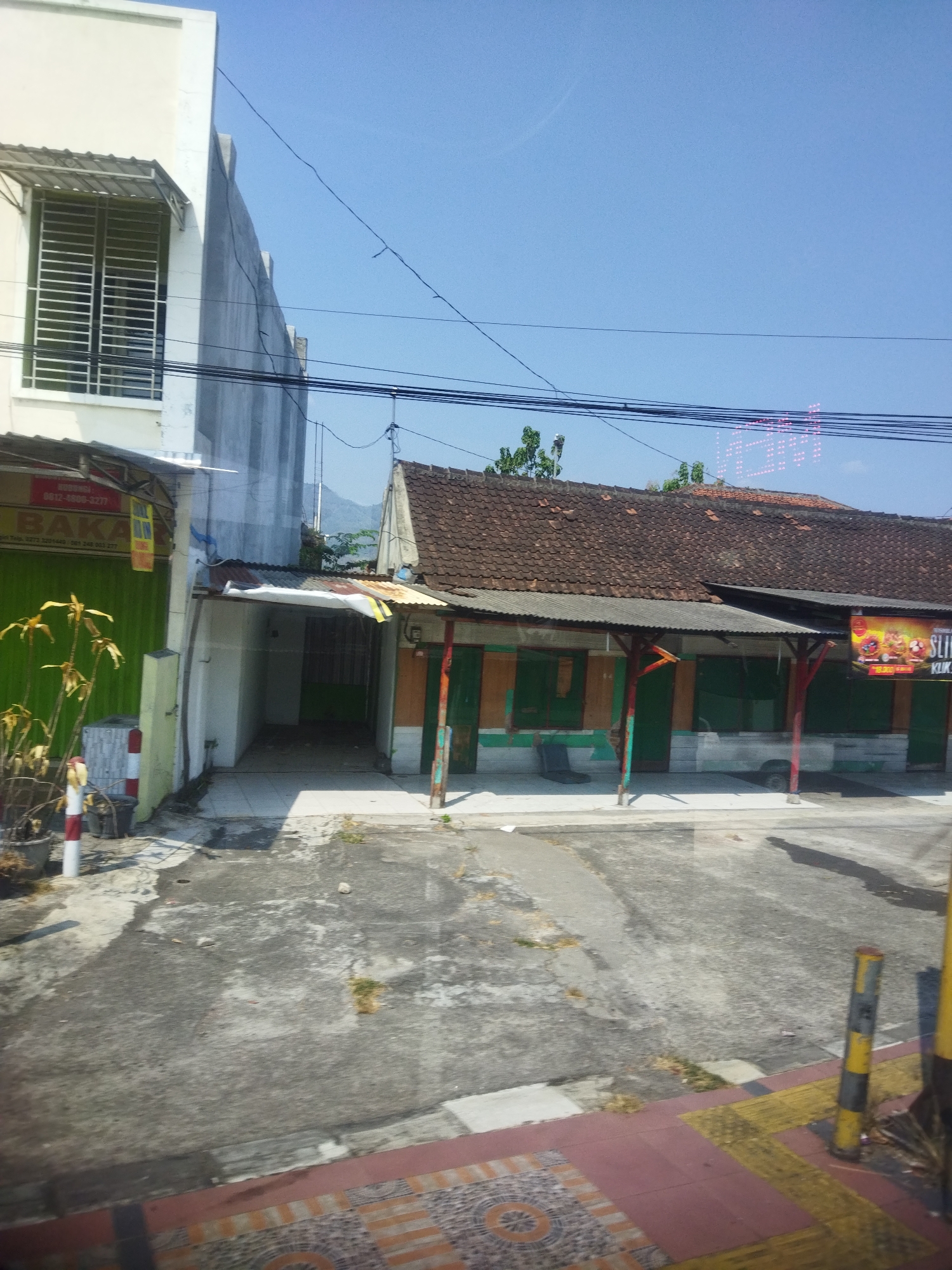 The image depicts a street scene in Singodutan, Indonesia, under a clear blue sky. On the left, a modern, multi-story building with white siding and a metal-grated window stands next to a narrower, older building with a green roll-up door. A red and white striped pole is positioned in front of the green door. Adjacent to this is a long, single-story structure with a brown tiled roof, supported by red and green wooden posts. This building has several green shuttered windows and a small awning. A sign with images of food items and text reading "SLIKI KLIK" is attached to the right side of this structure. Further back, mountains are visible in the distance. The street in the foreground is paved with asphalt and shows signs of wear and tear, with scattered debris and patches of dry vegetation. The right side of the image features a yellow and black striped pole and a patterned tiled walkway. The overall impression is that of a commercial area in a warm climate.
