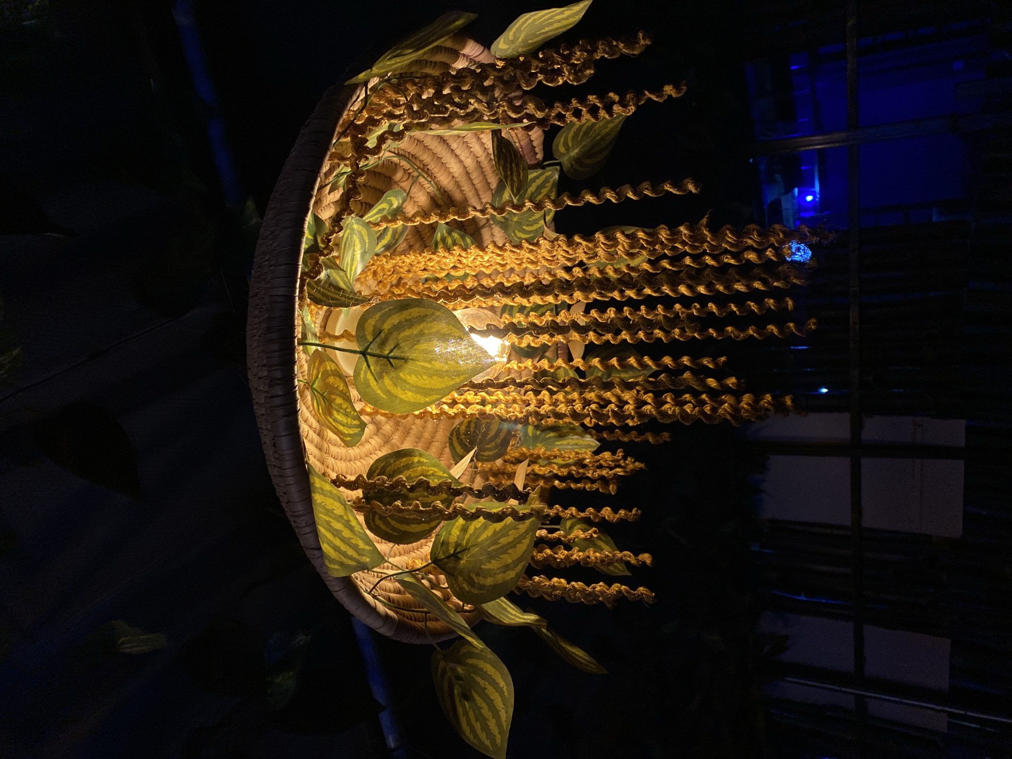 The image shows a decorative hanging lamp in Gura Top, Nigeria. The lamp is made from woven straw and decorated with artificial green leaves and long strands of yellow rope that hang down. The light bulb is visible inside the straw shade. The background is dark, suggesting it is nighttime or indoors. Faint blue lights are visible in the distance.