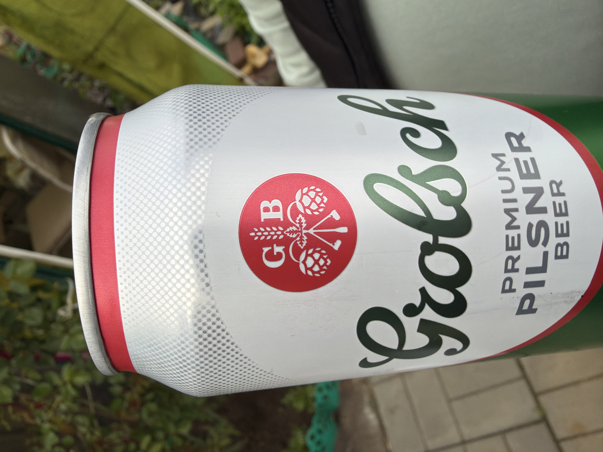 The image shows a close-up of a can of Grolsch Premium Pilsner beer. The can is oriented horizontally, with the top edge facing the left side of the image. The top of the can has a silver rim with a red band underneath. The main body of the can is white with a pattern of small gray dots. The Grolsch logo, in dark green cursive lettering, is prominently displayed on the can. A red circular emblem featuring a stylized hop and wheat design with the letters "GB" is also visible. Below the logo, the words "PREMIUM PILSNER BEER" are printed in black, block letters.
The background is blurred and suggests an outdoor setting with greenery. The exact location is not identifiable from the image.