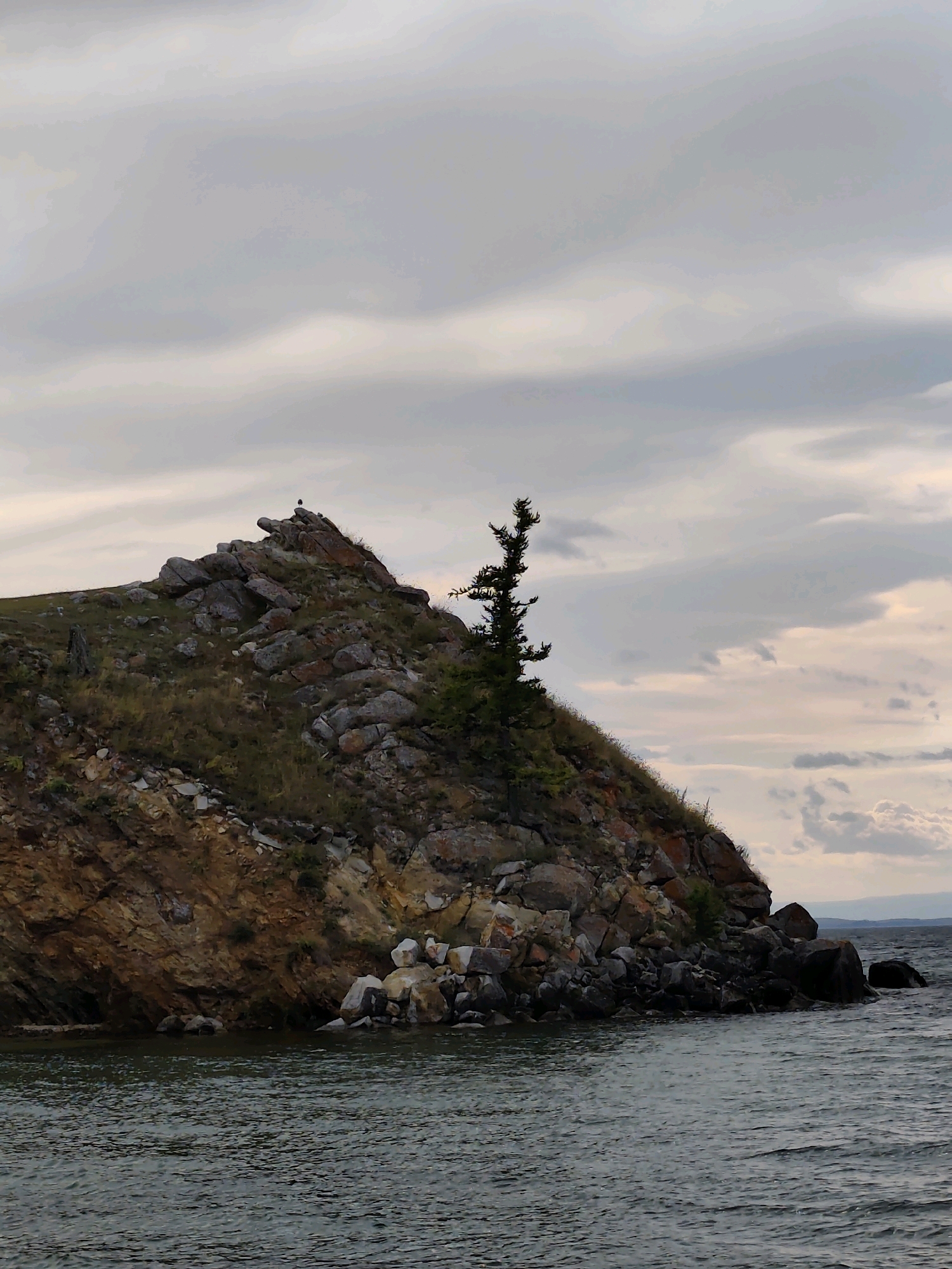 The image depicts a rocky island with sparse vegetation in the waters of Lake Baikal, near Kharantsy, Russia. The island is characterized by its rugged terrain, with large, irregularly shaped rocks scattered across its surface. A lone, slender tree with dark green foliage grows on the slope of the island, adding a touch of life to the otherwise barren landscape. The sky above is overcast with soft, diffused light, suggesting a cloudy day. The water of the lake appears dark and choppy, with small waves lapping against the shore of the island. In the distance, the faint outline of land can be seen on the horizon, shrouded in a light haze. The overall scene conveys a sense of rugged natural beauty and solitude.