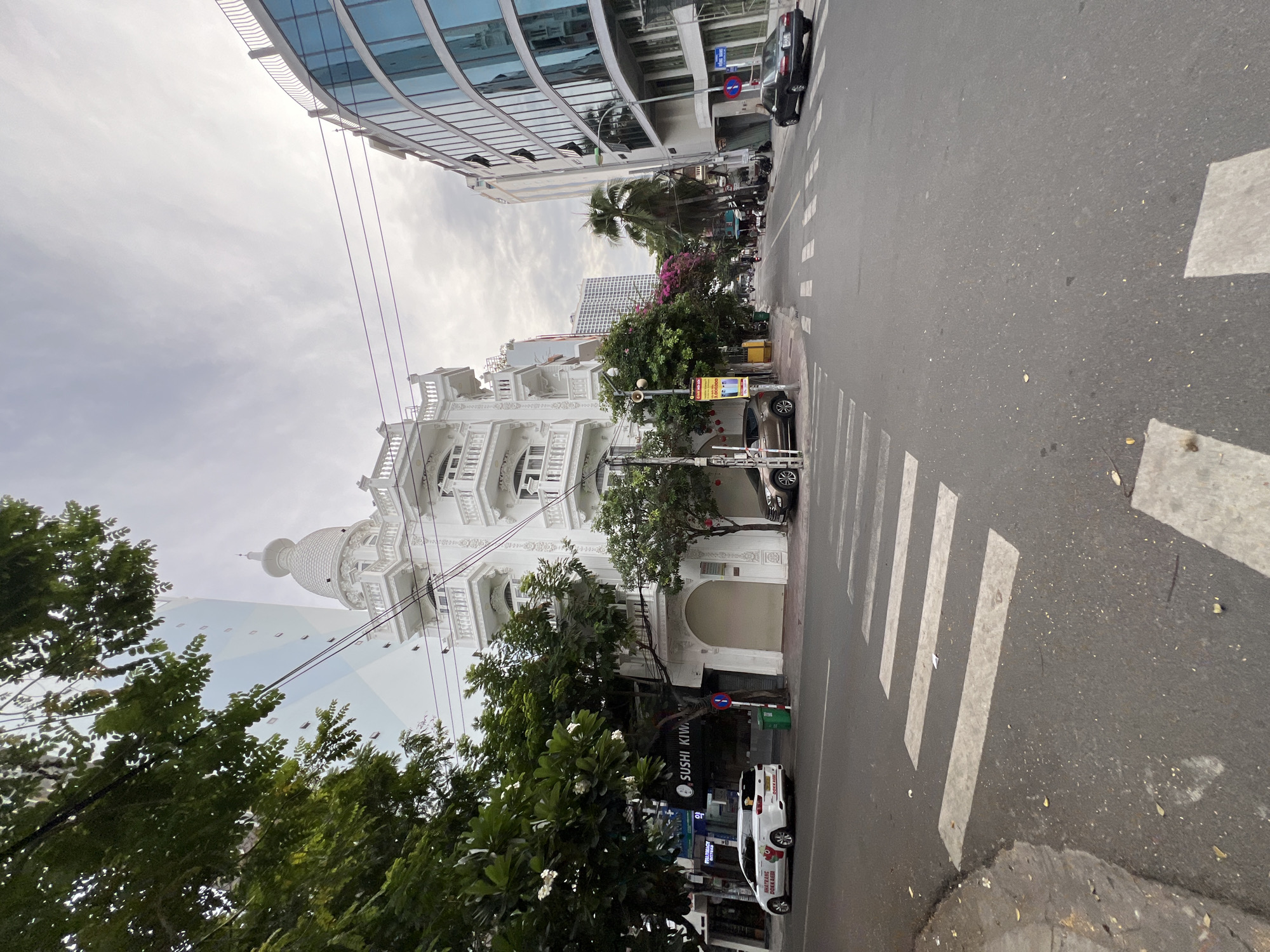 The image captures a street scene in Nha Trang, Vietnam, likely on a cloudy day. The foreground features a crosswalk marked with white stripes on an asphalt road. To the left, a white van with Vietnamese license plates and "HOANG TRANG DOKKAEBI" text is parked near a sidewalk. A white building with intricate architectural details, including balconies and a dome, stands prominently on the left side of the street, featuring a green trash bin and a red and white street sign. Adjacent to this building, on the left side of the street, is a business with a sign that reads "SUSHI KIWI".
To the right of the street, a modern building with a curved facade of blue glass windows dominates the landscape. Further down the street, other buildings and palm trees are visible. The sky is overcast with a few wispy clouds. The overall impression is of a busy urban environment with a mix of old and new architecture.