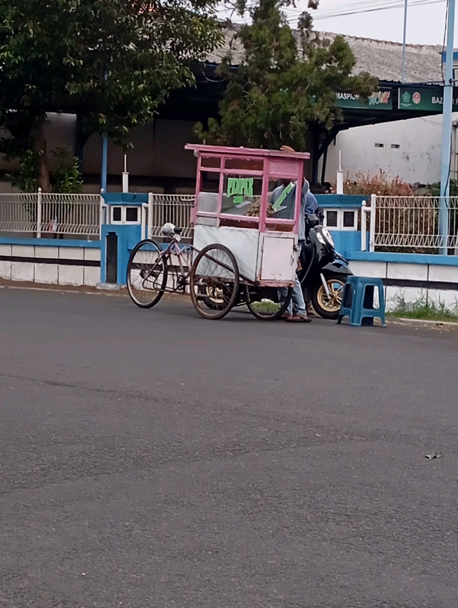 A stationary food cart, likely selling pempek, is parked on the side of an asphalt road in Randegan, Indonesia. The cart has a pink frame and a white body with the word "PEMPEK" visible in green lettering. A person, wearing jeans and a blue hooded jacket, stands beside the cart. To the right of the cart is a black scooter with gold-colored wheels and a blue plastic stool. In the background, a white building with a tiled roof is visible, along with trees and a white fence with blue accents. The sky is overcast.