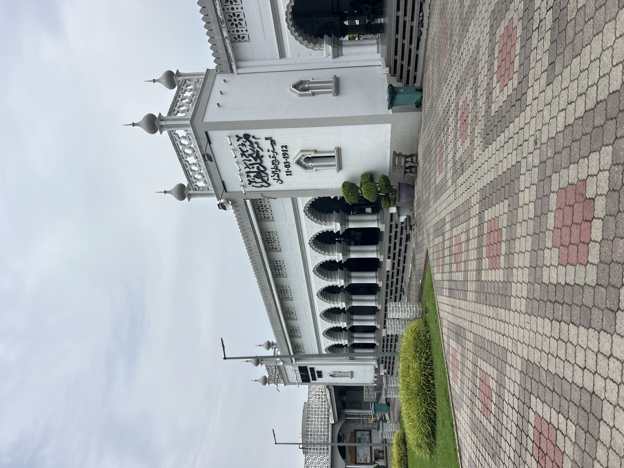 This is a medium-shot, eye-level, outdoor, daytime photograph of the Zahir Mosque in Alor Setar, Malaysia. The mosque is a large, white, Islamic religious building with a prominent tower. The architecture features arched walkways supported by columns, decorative latticework, and small domed finials on the tower. Arabic calligraphy and a date, "11-03-1912", are visible on the facade. In front of the mosque is a paved walkway with a distinctive pattern of interlocking grey and red tiles. To the left, there is a well-maintained lawn with manicured shrubs. A wooden bench and a potted plant are situated near the entrance. A green trash bin is visible to the right of the entrance. The sky is overcast and light grey, indicating a cloudy day. There are no discernible people in the immediate foreground, though shadows and faint figures suggest presence within the arched walkway of the mosque. The scene appears to be a street-level view of the mosque and its immediate surroundings.
