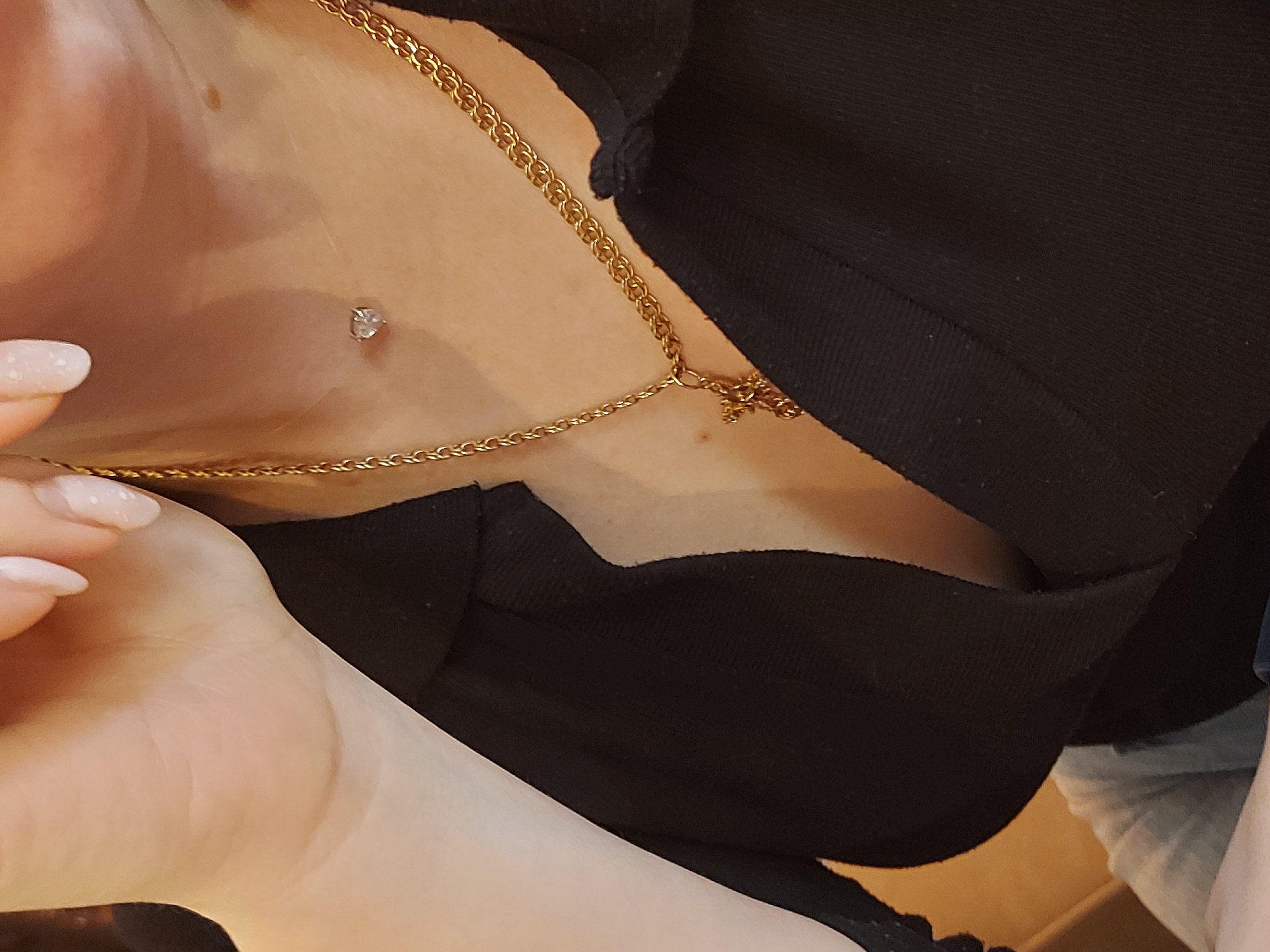 The image depicts a close-up view of an individual's upper torso and neck, captured in Chaikovsky, Russia. The subject wears a black collared garment, partially open at the neckline. A golden chain necklace with a small golden cross pendant rests on the upper chest. A dermal piercing, featuring a small, clear, heart-shaped gem, is present on the upper chest/lower neck area, positioned to the left. A portion of the individual's hand, displaying manicured fingernails with light-colored polish and white speckles, is visible at the upper left of the frame, near the neck. The skin tone is light, and minor dermatological features are present on the neck. The background is out of focus, revealing indistinct warm-toned elements, suggestive of an indoor environment, and a section of light grey fabric beneath the black garment.
