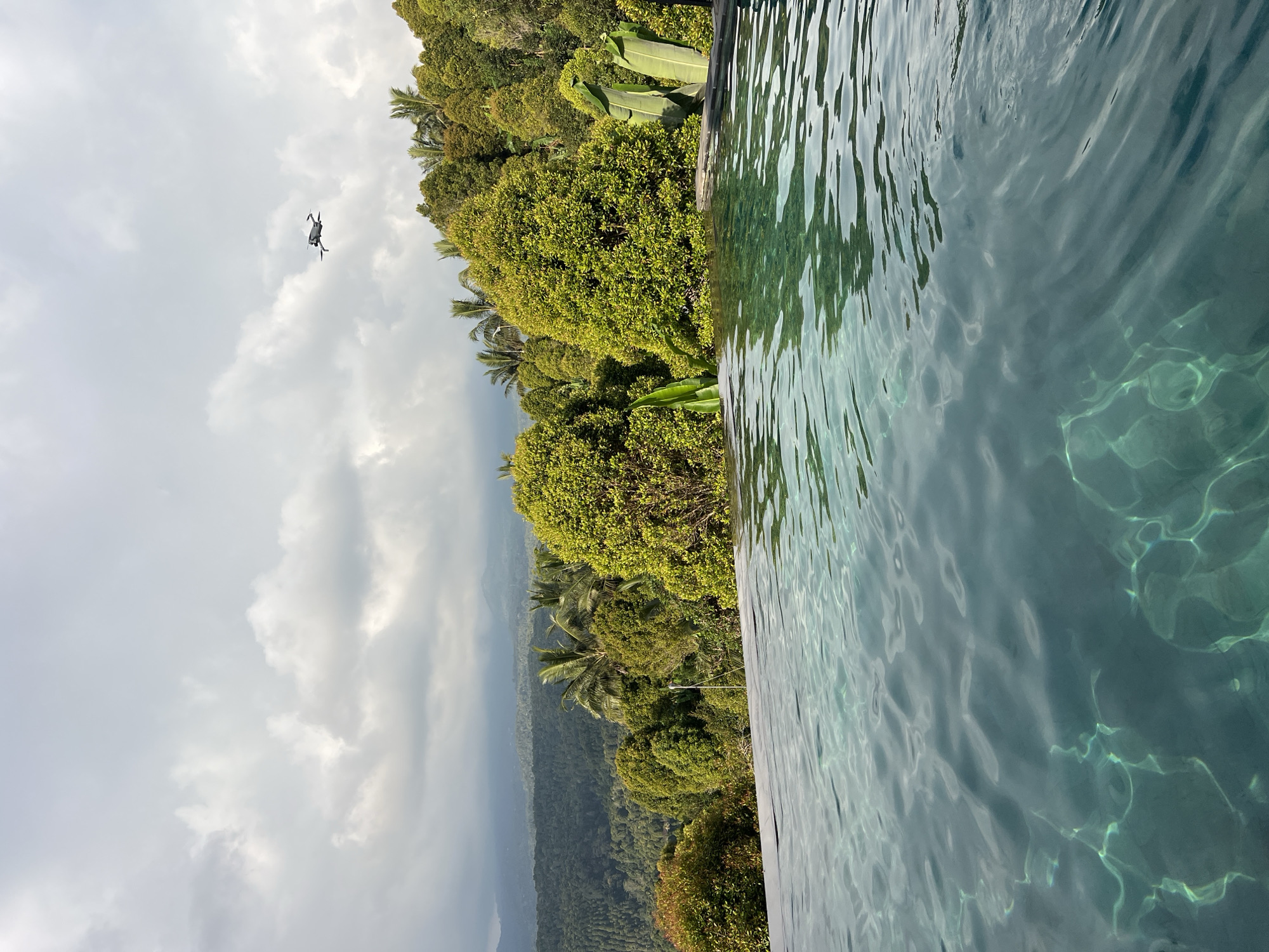 A drone flies in the distance above a lush, green landscape with rolling hills and dense trees. In the foreground, the edge of a swimming pool is visible, with clear blue water reflecting the sky and surrounding foliage. The sky is overcast with scattered clouds. The location is the city of Busungbiu, Indonesia.