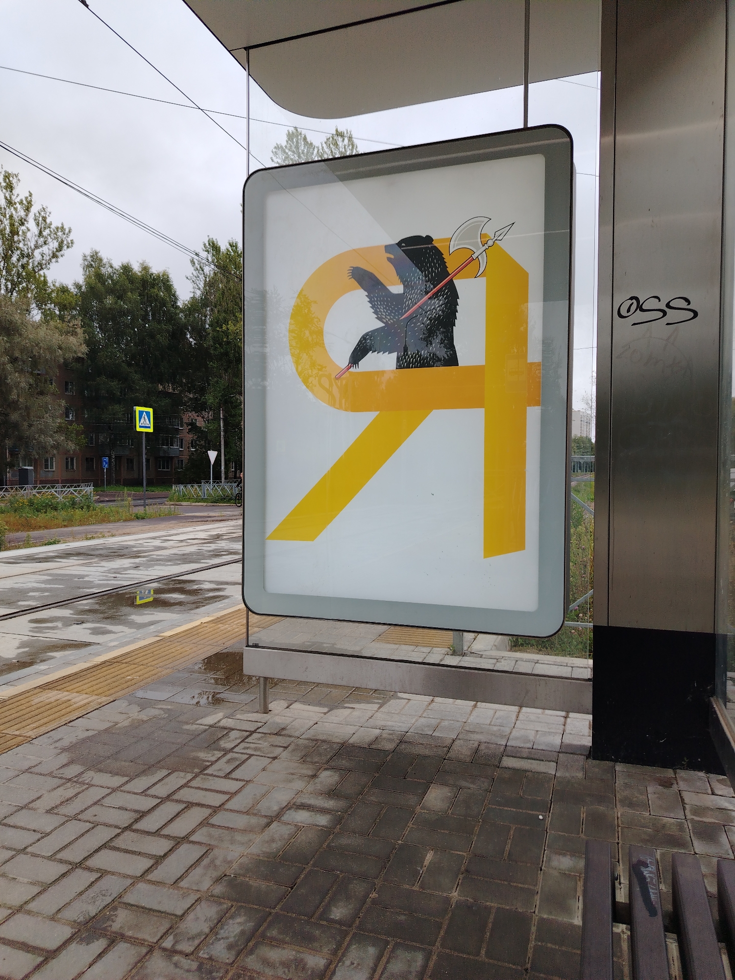 The image shows a close-up view of a billboard at a tram stop in Yaroslavl, Russia. The billboard features a stylized yellow "Я" with a black bear holding an axe. The bear is depicted in a dark, textured style, with its fur appearing bristly. The axe has a silver head and a red handle. The billboard is illuminated from behind, giving it a bright glow. To the right of the billboard, there is a graffiti tag that reads "OSS".
The tram stop itself is made of concrete pavers, which are wet, indicating recent rain. A yellow tactile paving strip runs along the edge of the platform. In the background, there are tram tracks, trees, and apartment buildings, suggesting an urban environment. The sky is overcast. There are no people clearly visible in the image, but the presence of a tram stop and a bench implies a public transportation area. The overall impression is that of a slightly damp and cloudy day in a Russian city.