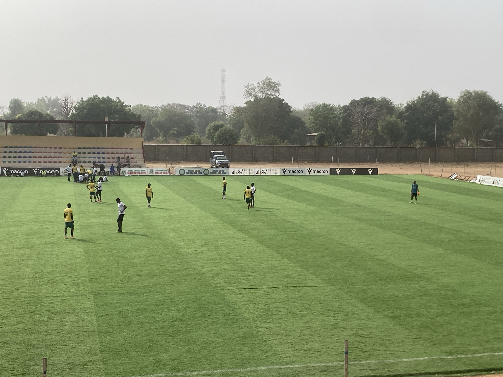 The image depicts a soccer field in Tudun Wada, Nigeria, likely a practice session. Several players in yellow jerseys and at least one in a blue jersey are dispersed across the green field. There are also a few individuals in white shirts. A small, partially filled stadium stands to the left of the field, and a vehicle is parked behind a wall, visible above banners with the "Macron" logo. Trees are in the background, and a telecommunications tower is visible behind the trees. The sky is hazy and overcast.