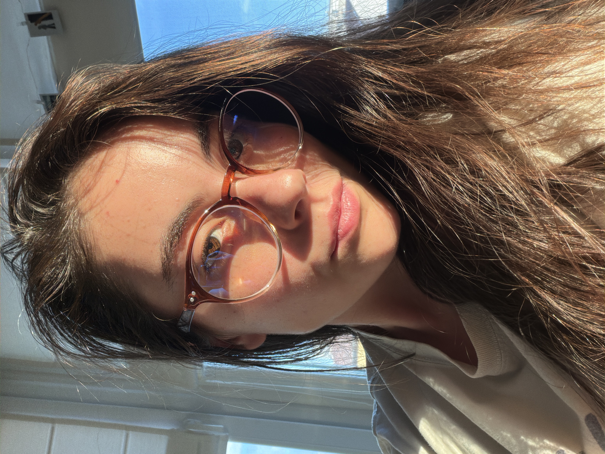 A close-up, tilted portrait of a young woman. She has long, brown hair and wears round, wire-rimmed glasses. Sunlight illuminates part of her face and hair. Her expression is neutral. The background shows a glimpse of a bright, sunny sky and a window frame. The location is Unknown, Unknown.