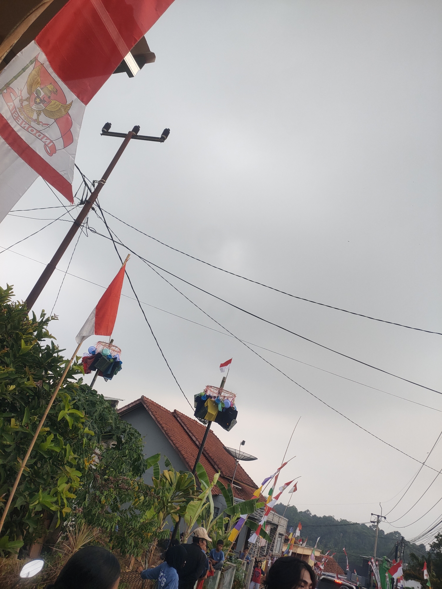 The image depicts a street scene in Subang, Indonesia, likely during a celebration or national holiday given the prominent display of Indonesian flags. The flags are red and white, with one large flag in the foreground featuring the Garuda Pancasila emblem. Several poles with flags and decorative elements are visible along the street. Overhead power lines crisscross the sky, which is overcast. In the background, residential buildings with red tile roofs are present, along with lush green foliage and banana trees. Several people are visible, some standing and others possibly walking or sitting. The overall impression is a vibrant community event.