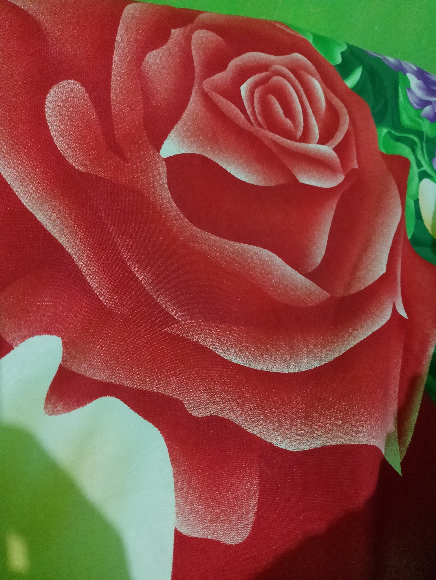 A close-up, top-down view of a red rose pattern on fabric. The rose is the dominant feature, with layers of petals depicted in shades of red, white, and light pink, creating a sense of depth and texture. The fabric appears to be textured, with a subtle weave visible. In the upper right corner, there are hints of green leaves and purple flowers, suggesting a floral bouquet or garden theme. The background transitions from a bright green at the top to a darker red at the bottom, with a soft shadow cast on the lower right section, indicating artificial lighting. There are no people or identifiable landmarks present that would allow for specific location identification in Rembang, Indonesia.