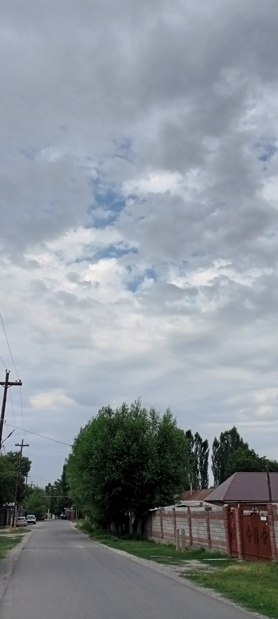 The image depicts a street scene in Sokuluk, Kyrgyzstan. The perspective is from a low angle, looking down a paved road. The sky is overcast with numerous grey and white clouds, with patches of blue sky visible in the upper portion of the frame. On the left side of the road, a utility pole with wires runs parallel to the street. Further down the road on the left, several parked cars are visible. The right side of the road is lined with a brick and stone fence, topped with a concrete cap. Beyond the fence, parts of a building with a dark roof and decorative wrought iron gate are visible. A large, lush green tree dominates the middle ground, with a line of taller, slender trees behind it. The ground on both sides of the road appears to be grass and dirt. The overall impression is of a quiet, residential street in a town or village.