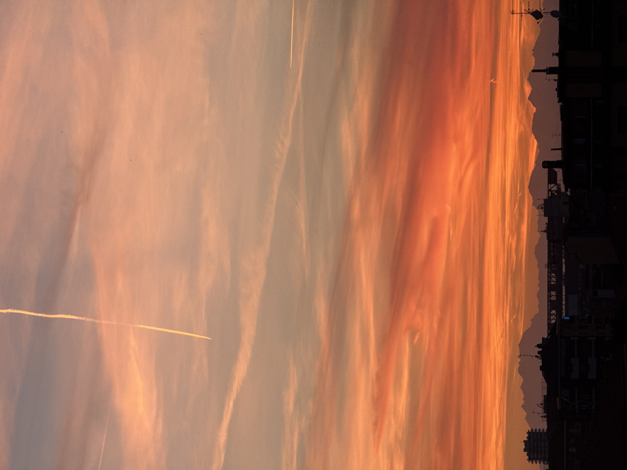 The image shows a vertical, slightly rotated view of a sunset over Milan, Italy. The sky displays vibrant hues of orange, peach, and pale blue. There are contrails of airplanes cutting across the sky. The silhouette of the city skyline is visible at the bottom, with building rooftops, antennas, and distant hills and mountains. The lighting indicates a sunset, casting the buildings into dark silhouette. The image quality suggests it was taken from a high vantage point. No people are visible.