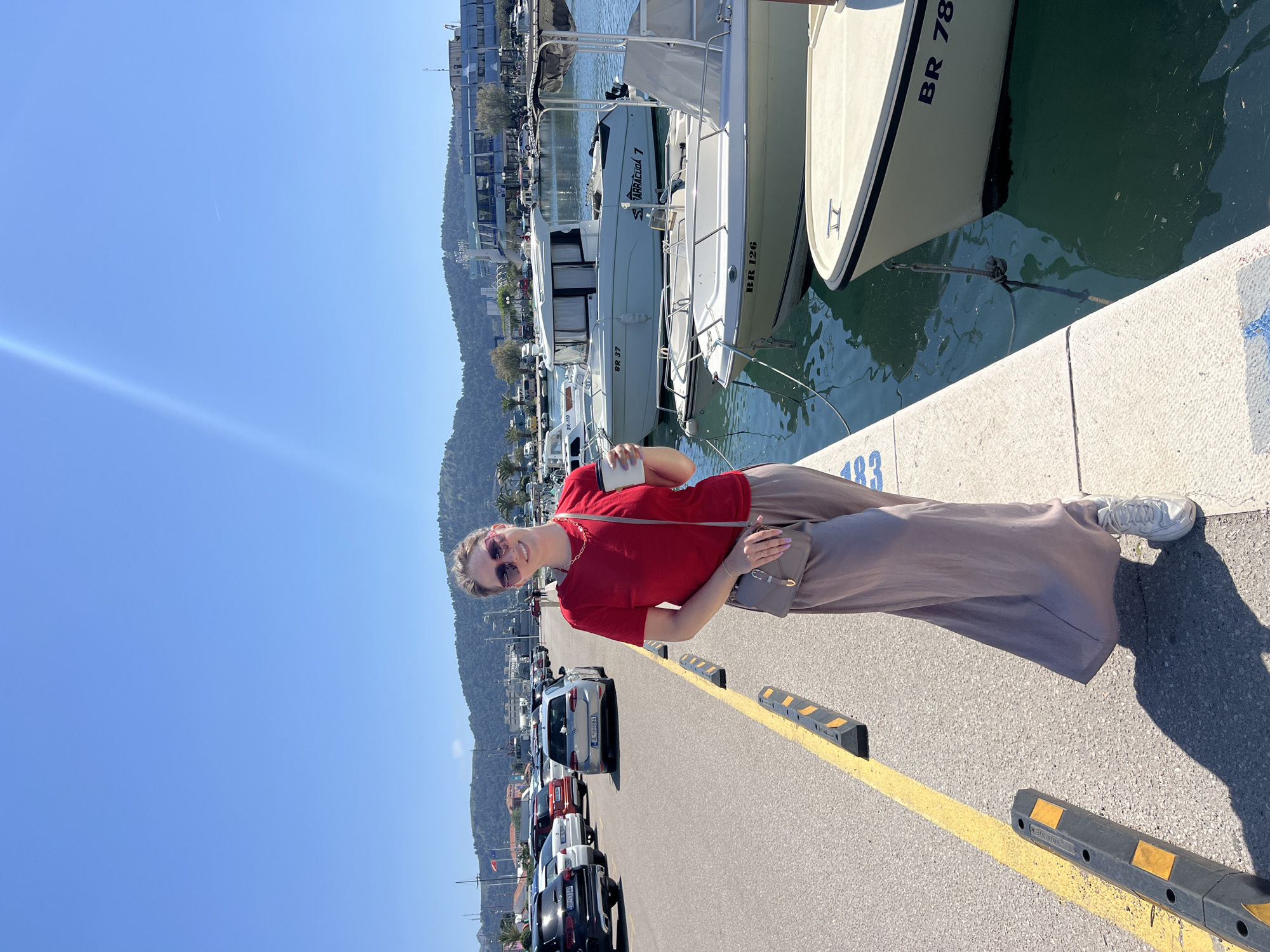A woman stands on a concrete pier next to docked boats in Bar, Montenegro. The woman wears a red t-shirt, taupe pants, white sneakers, and sunglasses, holding a cup and a small purse. Behind her are rows of parked cars, a yellow line marking the edge of the pier, and a mountain range in the background. Boats are moored along the pier. The sky is clear and blue. The number "483" is painted on the edge of the pier. Buildings and trees are visible in the distance.