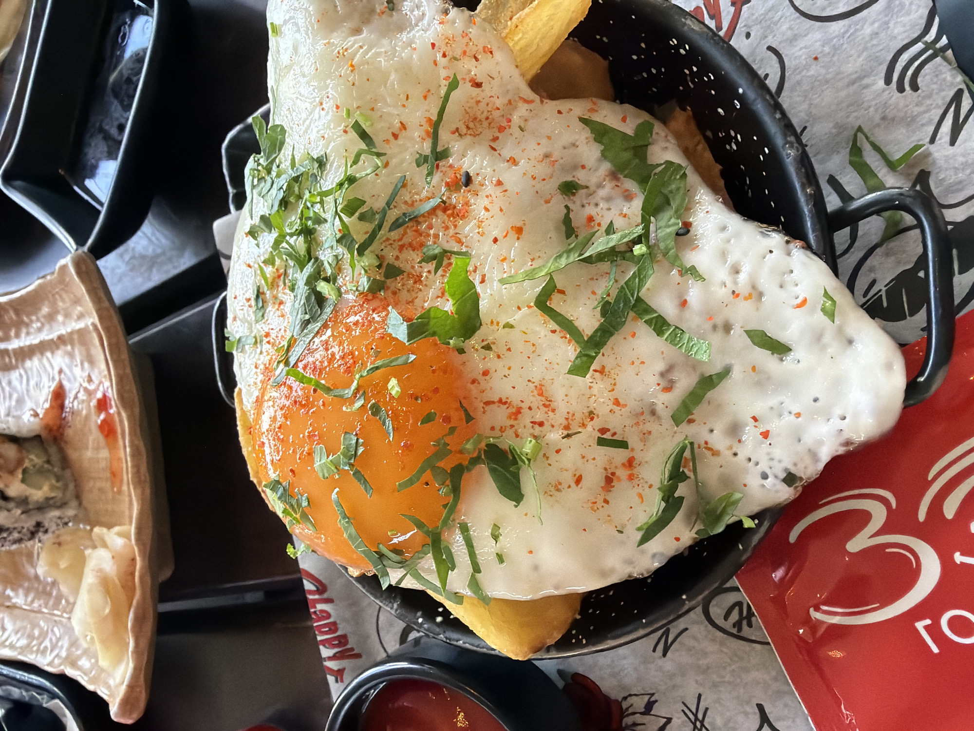 The image shows a close-up of a black metal basket containing fries topped with a fried egg, sprinkled with chopped green herbs and reddish-orange powder. The egg has a bright orange yolk. Next to the basket is a small bowl containing ketchup. Part of a plate with sushi or other food items is visible to the left. The setting appears to be a restaurant or cafe, as suggested by the patterned paper underneath the food and the presence of other dishes nearby. A red packet with white lettering (partially visible) indicates the location is likely in Varna, Bulgaria, due to the use of Cyrillic script "ГОД." appearing on the packet.