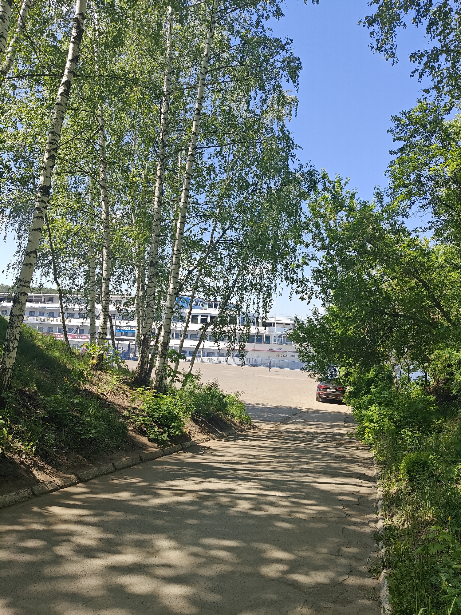 The image depicts a downward sloping paved road in the city of Chaikovsky, Russia. The road is flanked by trees and vegetation on both sides, with birch trees prominent on the left. Sunlight filters through the trees, casting shadows on the road. In the distance, a multi-decked river cruise ship is visible along the waterfront. A dark-colored car is parked on the road further down, near the tree line on the right. The sky is clear and blue.