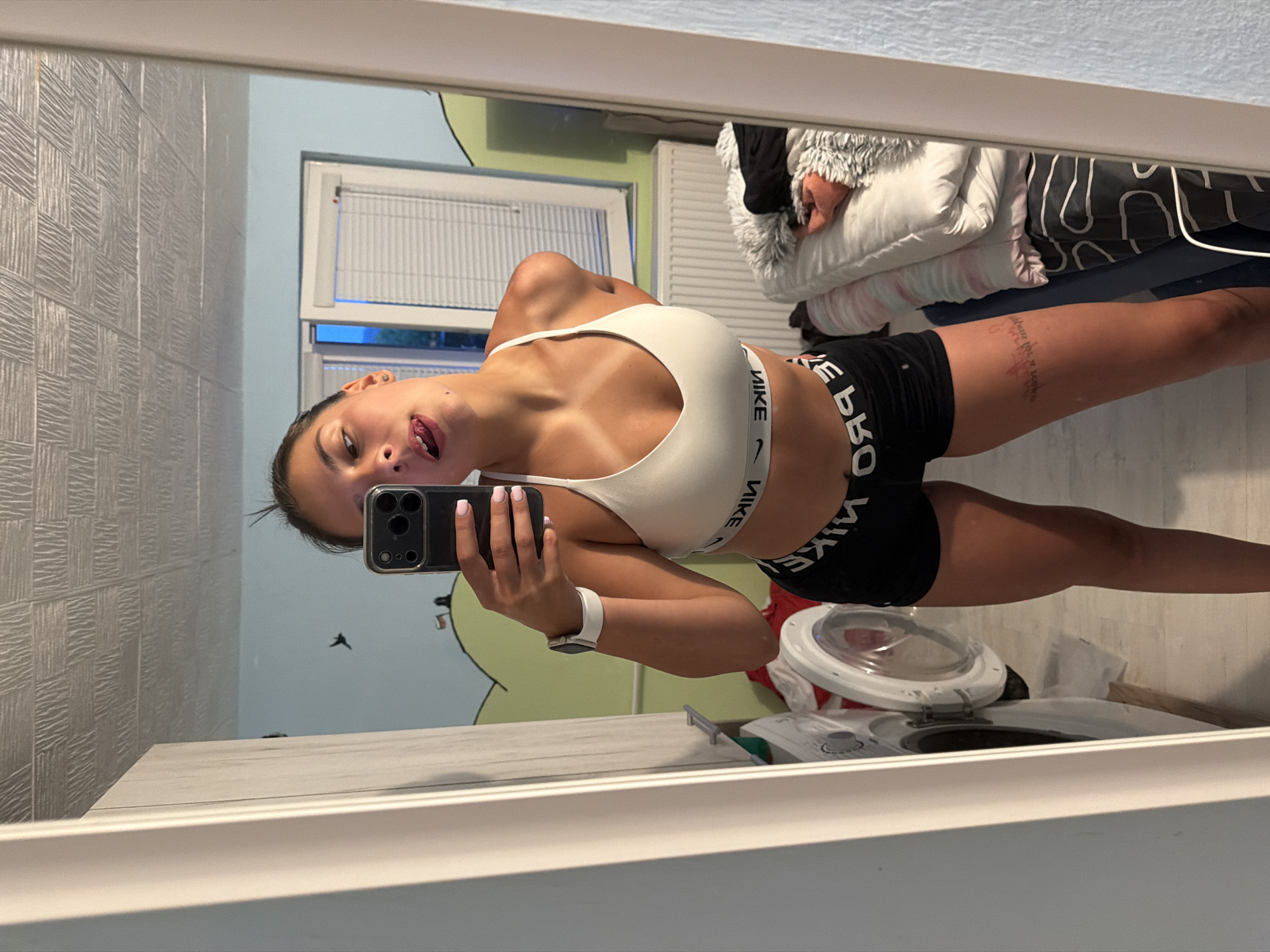 In a room, a young woman is taking a mirror selfie. She is wearing a white Nike sports bra and black Nike shorts. She has a smartwatch on her left wrist. Her hair is pulled back in a ponytail, and she is sticking her tongue out at the camera. The room appears to be a bedroom or a dressing area, with a washing machine visible to the left and a bed with bedding to the right. The walls are decorated with what appears to be a mural of hills and a sky. The ceiling is textured. The image is a snapshot of a personal moment, captured in an indoor setting.