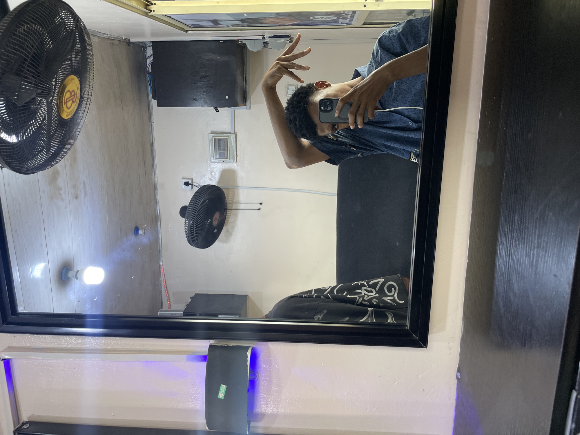 A person with dark skin and short, dark curly hair, wearing a blue patterned shirt, is holding a smartphone and posing with their hand up in the air, reflected in a large rectangular mirror with a black frame. The reflection shows the person standing near a black couch with a patterned throw, in a room with white walls. Visible in the reflection are two electric fans, one large and one smaller, a light fixture, and what appears to be a black electronic device mounted on the wall. There is also a black object with a blue light attached to the bottom of the mirror frame. The image was taken in Ado, Nigeria.
