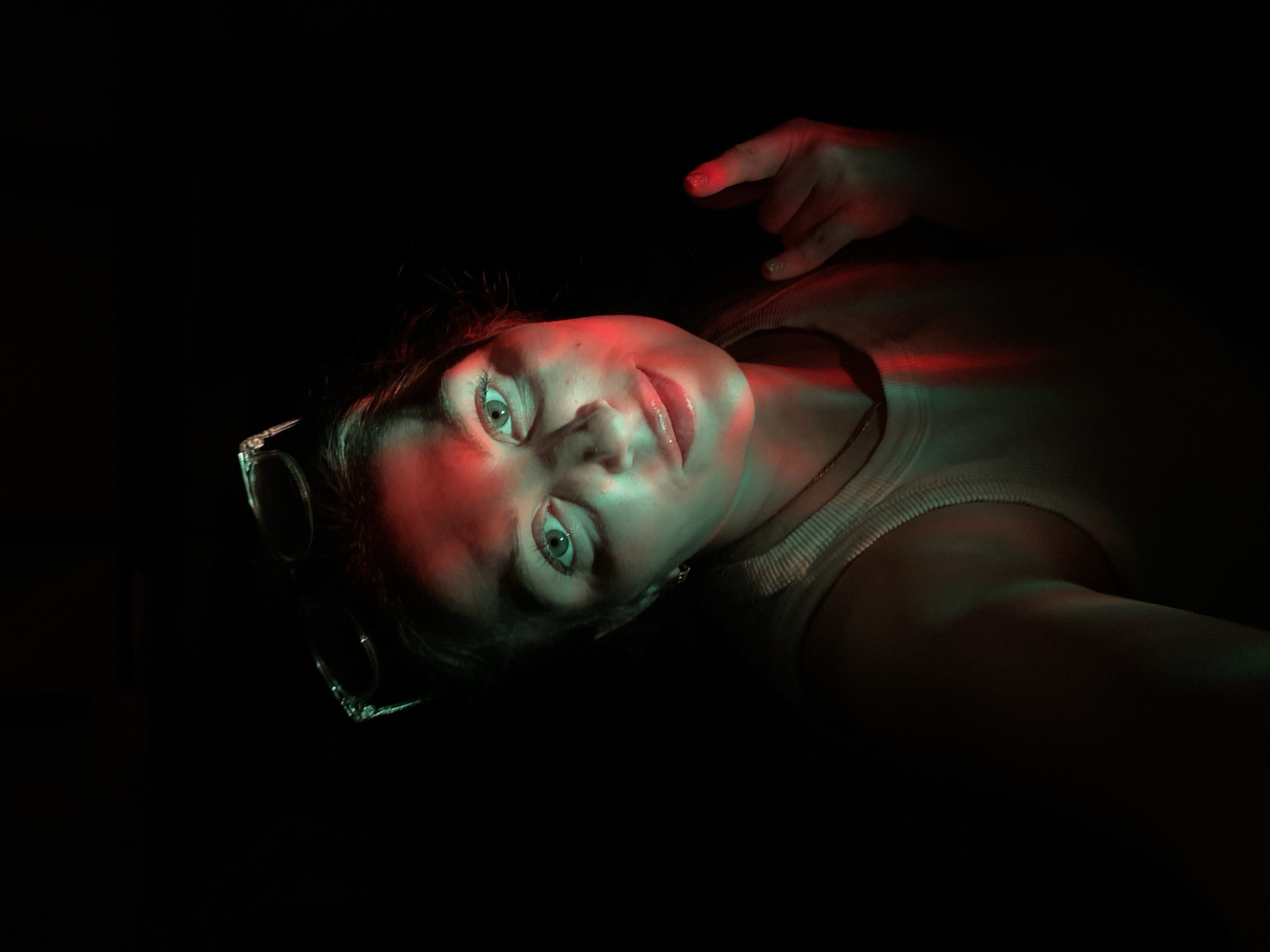 A woman is lying down in near darkness, illuminated by red and green light. Her face is turned towards the camera, with her hand raised and pointing a finger. She wears a light-colored tank top. Sunglasses are on her head. The location in the city of Марковский, Russia, cannot be determined from this image.