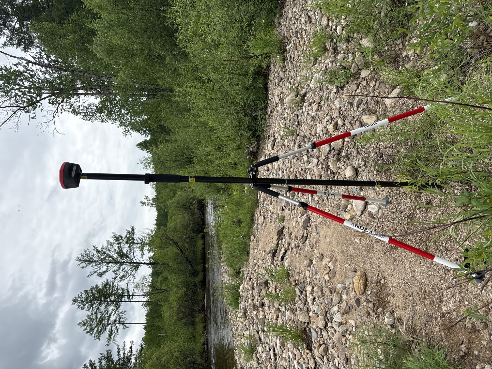 An outdoor, eye-level shot captures a surveying instrument, likely a GPS receiver, mounted on a tripod in the foreground. The instrument is positioned on a rocky, gravelly riverbank with scattered green vegetation. Behind the instrument, a body of water, a river, flows gently. The background is dominated by lush green trees and foliage, with a cloudy, overcast sky above. The location is identified as Murtygit, Russia. The scene is static and shows no people or any ongoing activity.