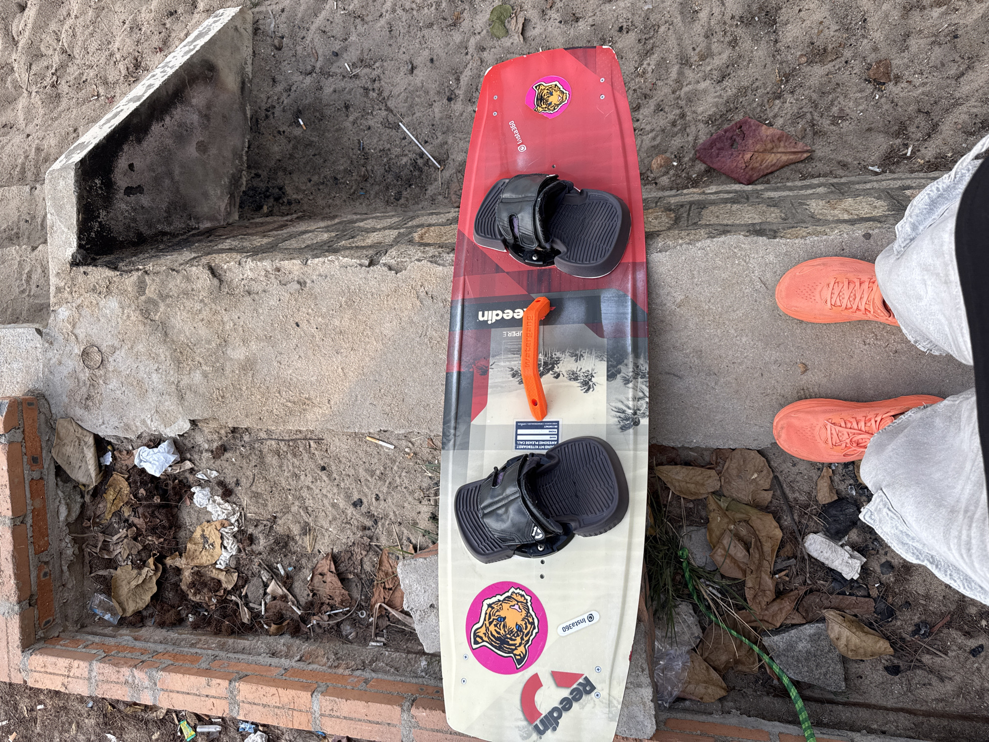 The image depicts a wakeboard with orange bindings placed on a sandy surface adjacent to a concrete structure. The wakeboard is oriented horizontally and shows a tiger sticker on its surface. In the background, a brick structure filled with debris, including dry leaves, plastic, and cigarette butts, is visible. The scene appears to be outdoors, possibly in a less-maintained area of Nha Trang, Vietnam, given the sand and weathered concrete. The presence of what appears to be a wakeboard suggests a watersport-related location.