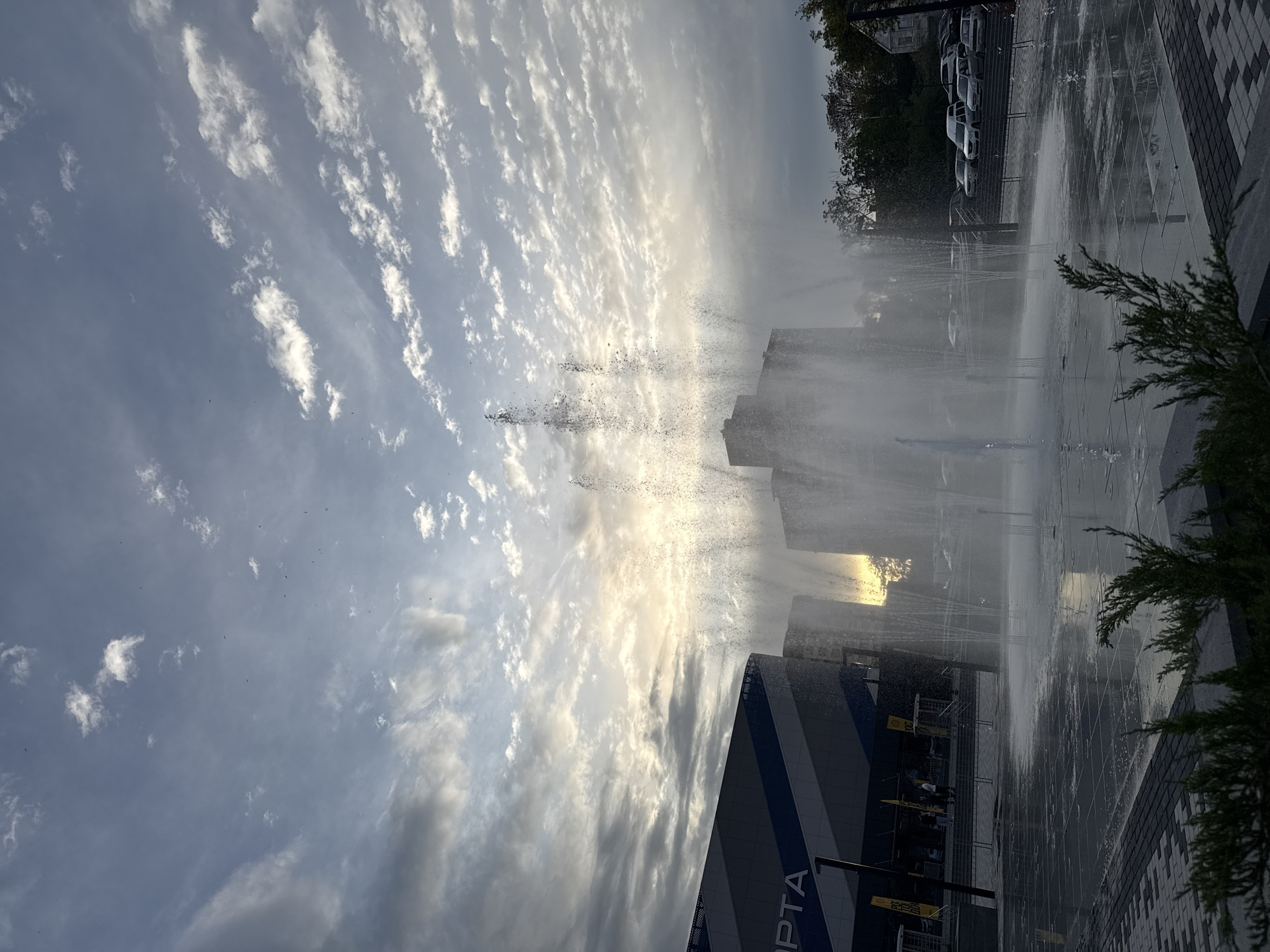 The image depicts a water fountain in a public space in Rostov-on-Don, Russia. The fountain sprays water vertically into the air. A building with the inscription "ROSTA" is visible on the left side of the fountain. The sky is cloudy with bright sunlight filtering through the water streams. A sidewalk borders the fountain on the right with trees on the top right. Parked cars are seen in the background.