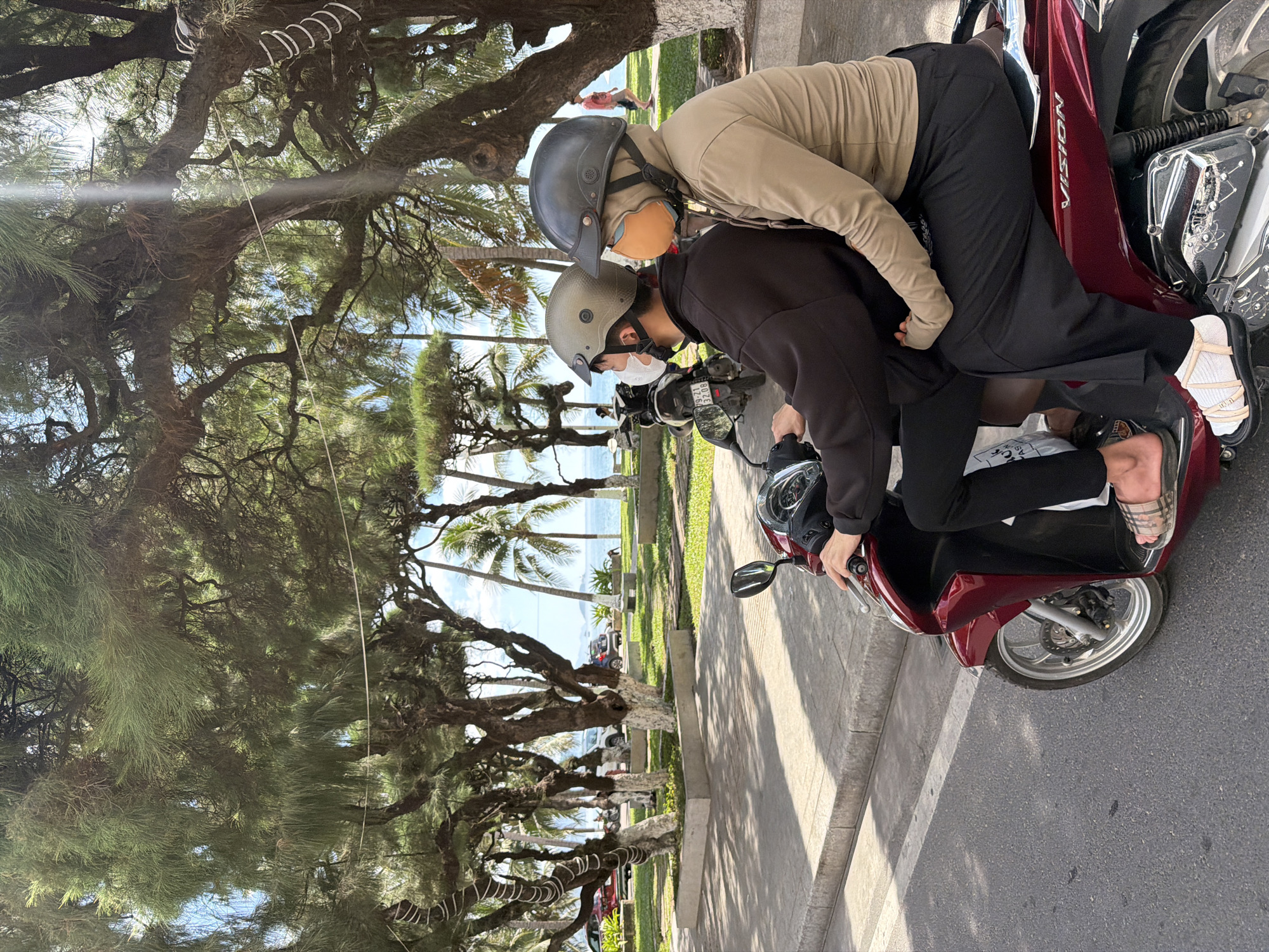 Two individuals, both wearing helmets and face masks, are riding on a maroon scooter. The driver, in the front, wears a dark-colored jacket and black pants. The passenger, behind the driver, is wearing a tan-colored hoodie and black pants. They are on a paved road with a sidewalk and a grassy area to their right. Beyond the grassy area, there are trees, palm trees, and a body of water that appears to be the ocean, suggesting a coastal location. In the background, several cars are parked, and a person is walking. The sky is bright and appears to be daytime. This scene is consistent with a location in Nha Trang, Vietnam, known for its beaches and coastal scenery.