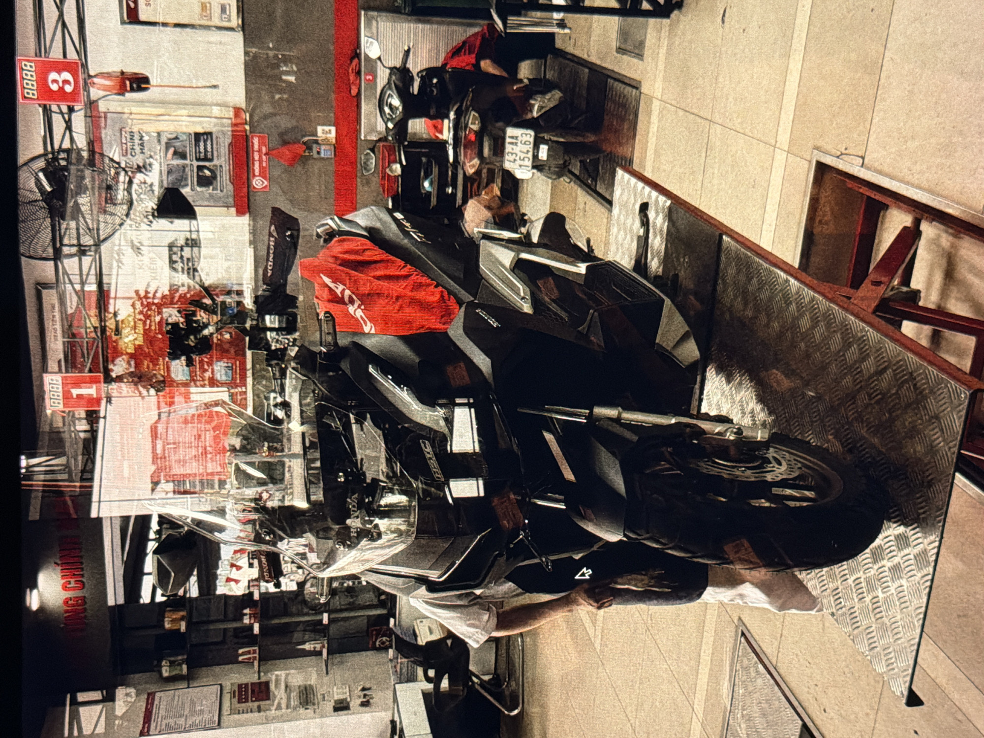 The image shows a Honda motorcycle repair shop in Đà Nẵng, Vietnam. Two Honda motorcycles are visible; the first is black and positioned on a diamond-plated platform. The second motorcycle is black, parked on the tiled floor nearby, and partially obscured. An individual in a white shirt is bent over, facing one of the motorcycles. Overhead, a red sign with white numerals displays "1" and "3" next to the shop's entrance. Another sign reads "HÔNG CHÍNH", and a third says "KIEM CHÍNH HÃN". The Vietnamese license plate of one of the motorcycles shows "43-AA 154.63". The environment includes shelves with various motorcycle parts and accessories.