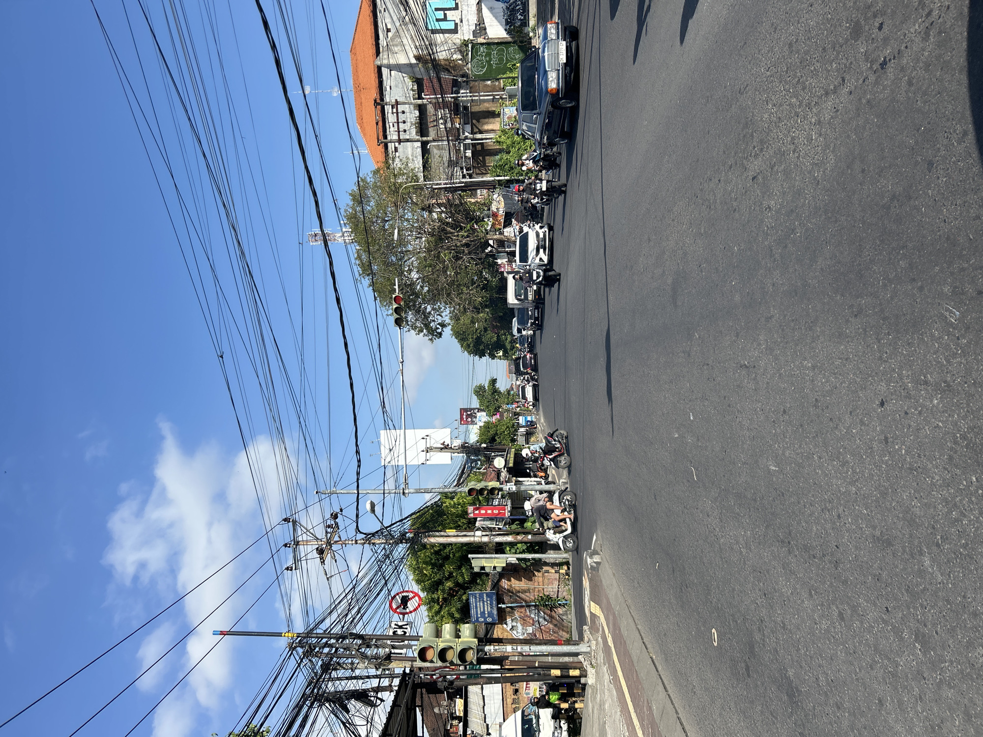 The image depicts a street scene in Denpasar, Indonesia, under a bright, partly cloudy sky. A paved road dominates the foreground, with a sidewalk to the left. Several vehicles, including cars and motorcycles, are present on the road, some in motion. Traffic lights and numerous utility poles with a dense network of electrical wires are prominent features, indicating a busy urban environment. Buildings line the street, with some featuring distinctive Balinese architectural elements like orange tiled roofs. People are visible riding motorcycles. The overall impression is a snapshot of everyday urban life in Denpasar, characterized by traffic, infrastructure, and architectural styles.