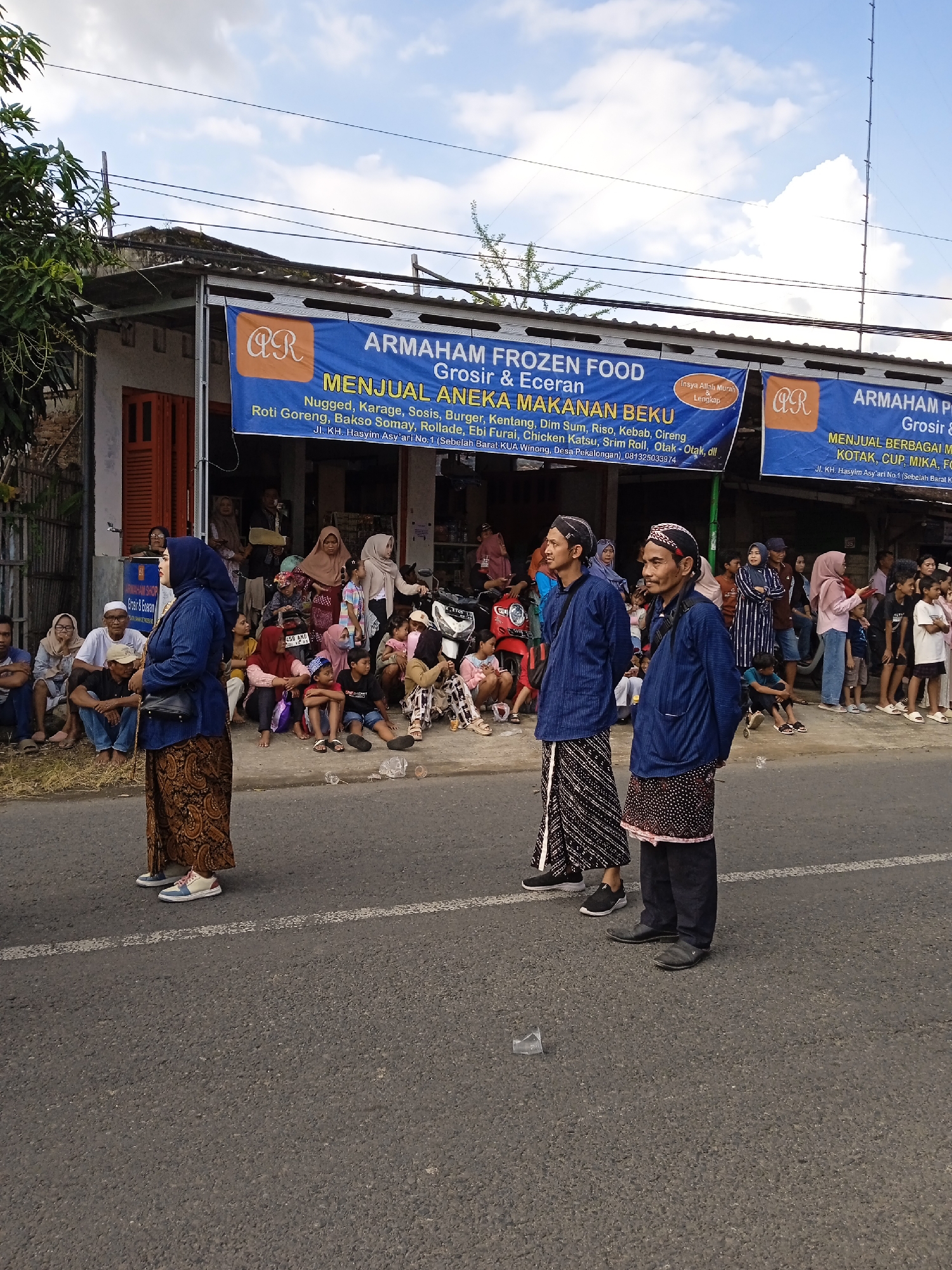 The image captures a street scene in Winong, Indonesia, in front of "ARMAHAM FROZEN FOOD," a shop selling frozen food and packaging materials. A crowd of people, including women in hijabs and children, are seated and standing along the side of the road, seemingly observing an event. In the foreground, three individuals are standing on the road. One is a woman wearing a headscarf and a traditional skirt. Two men are wearing traditional attire, including patterned sarongs and jackets with headbands. The asphalt road shows some litter. The sky is partly cloudy.